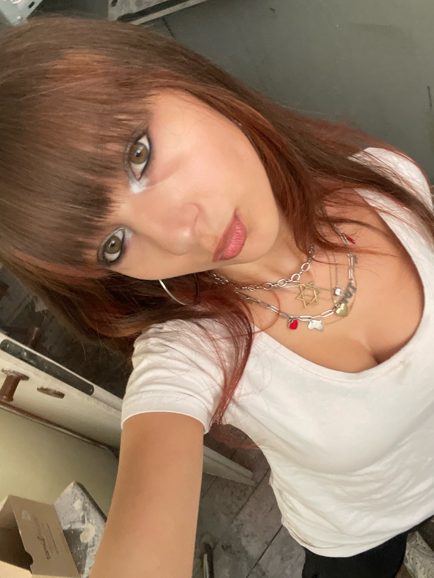 This is a close-up, angled selfie of a person with auburn hair and bangs, wearing a white t-shirt and dark pants. They have heavy eye makeup, including white eyeshadow. They are wearing multiple necklaces, one with a Star of David pendant, a heart pendant, and other charms. The background appears to be an indoor space with a rough-textured floor and some objects in the lower left corner that resemble construction materials or debris. The lighting is somewhat harsh, creating shadows. No specific location in Raanana, Israel is identifiable.