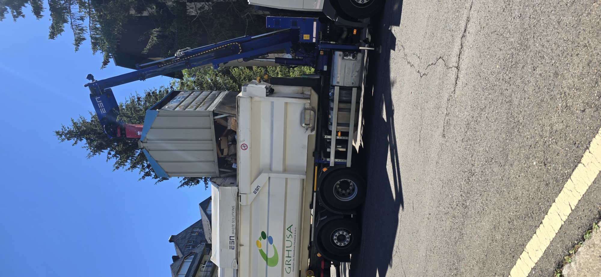 A blue crane arm, attached to a white garbage truck, is lifting a large, light-colored debris bin into the back of the truck. The bin appears to be filled with various types of refuse, including wood and cardboard. The truck has "ESA WASTE SOLUTIONS" and a green and yellow logo with the text "GRHUSA Gestión de Residuos Huesca s.a.u." printed on its side. The truck is parked on an asphalt road with a yellow line painted on the edge. In the background, there are green trees and the roof of a building. The sky is clear and blue, indicating a sunny day. The scene appears to be set in a residential or commercial area, possibly during a waste collection service. The image is oriented vertically.