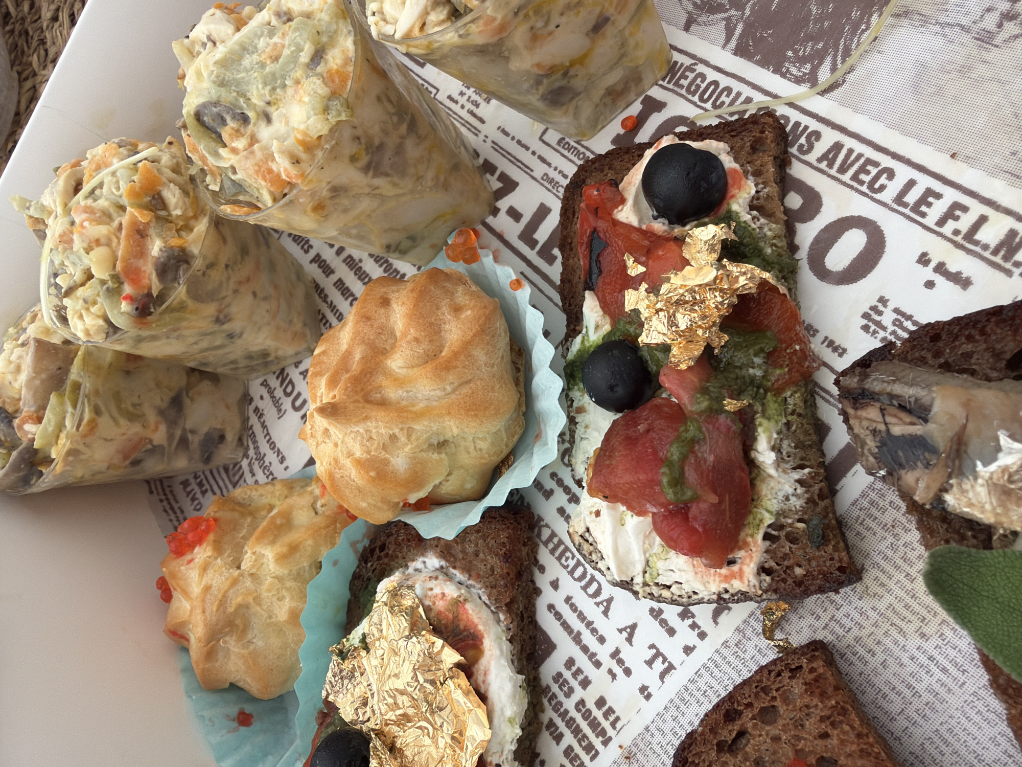 An overhead shot captures an assortment of meticulously prepared appetizers or amuse-bouches displayed on a white platter and a paper liner designed to resemble vintage French newspaper. The setting appears to be a casual yet upscale catering event or gathering in Bat Yam, Israel.
On the left, several clear plastic cones or cups are filled with a creamy, chunky salad, possibly a potato or vegetable salad with visible pieces of carrots, green elements, and possibly mushrooms, suggesting a rich, savory filling.
Towards the center and right, various open-faced canapés are presented on slices of dark, whole-grain bread. One prominent canapé features a white spread, topped with vibrant red roasted peppers, a black olive, a drizzle of green pesto or sauce, and luxurious gold leaf. Another partially visible canapé also includes gold leaf and a white spread. A separate piece of dark bread is topped with what appears to be a small fish, possibly a sardine.
Interspersed with the canapés are light golden choux pastry puffs, some placed in light blue fluted paper liners and decorated with small red elements. A large, fuzzy green leaf, possibly sage, is visible on the far right. The overall impression is one of elegant and diverse finger foods.
The paper liner beneath the food features text in French, designed to mimic an old newspaper. Visible phrases include "NÉGOCIONS AVEC LE F.L.N." (Negotiate with the F.L.N.), "EDITION," "DIREC," dates like "1943," and other fragmented words such as "KHEDDA A TI," "combats," and "BEN BEL," suggesting a historical or themed presentation. The lighting is bright and natural, indicating a daytime setting.