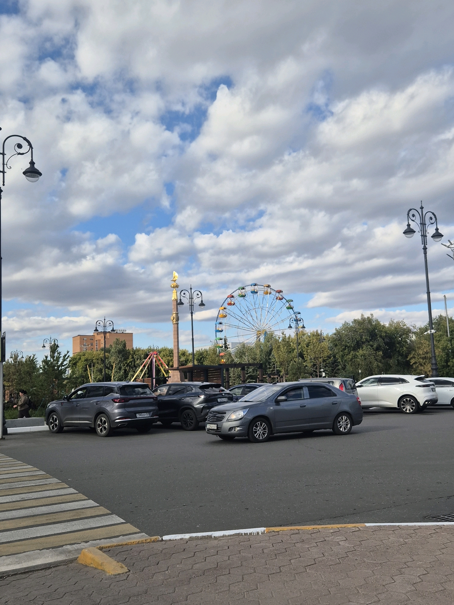 The image depicts a street scene in Kokshetau, Kazakhstan, featuring a road with several cars, a pedestrian crossing, and a distant view of an amusement park with a Ferris wheel and a monument. The sky is partly cloudy with a mix of white and gray clouds against a blue backdrop.
In the foreground, a pedestrian crossing with yellow and white stripes is visible, leading to a paved sidewalk. A yellow concrete curb stop is present near the crossing. The road surface is asphalt.
Several cars are present on the road. Closest to the foreground on the left is a dark gray SUV. To its right, a black SUV is parked. Further ahead and to the right, a gray sedan is in motion. In the background, a white crossover SUV is parked, with another white vehicle partially visible behind it.
The background shows trees, a building with windows, a golden monument topped with a decorative element, and an amusement park with a colorful Ferris wheel. Streetlights with a decorative design are also visible on poles. A person is seen walking on the sidewalk on the far left of the image. The overall impression is a bright, overcast day in an urban setting.