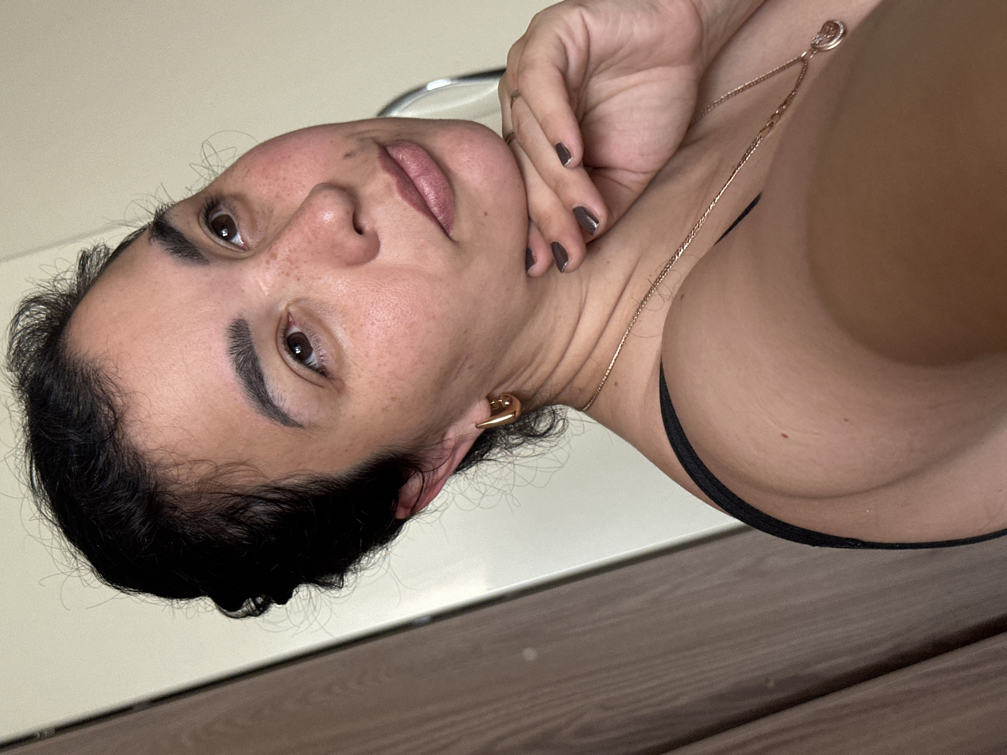 The image shows a close-up, vertically oriented portrait of a person, likely a woman, from the chest up. She has dark hair, dark eyebrows, and brown eyes. Her skin has a warm tone with visible freckles. She is wearing a black top and a rose gold necklace with a small pendant. A gold earring is visible in her left ear. Her right hand is resting on her face, near her cheek and chin, with dark nail polish on her fingernails. Her lips are painted with a matte lipstick. The background is a plain, light-colored wall, and the lower portion of the image shows a wooden surface. The lighting appears to be soft and natural, suggesting it could be daytime indoors. The person's expression is contemplative or relaxed. There is no visible text in the image.