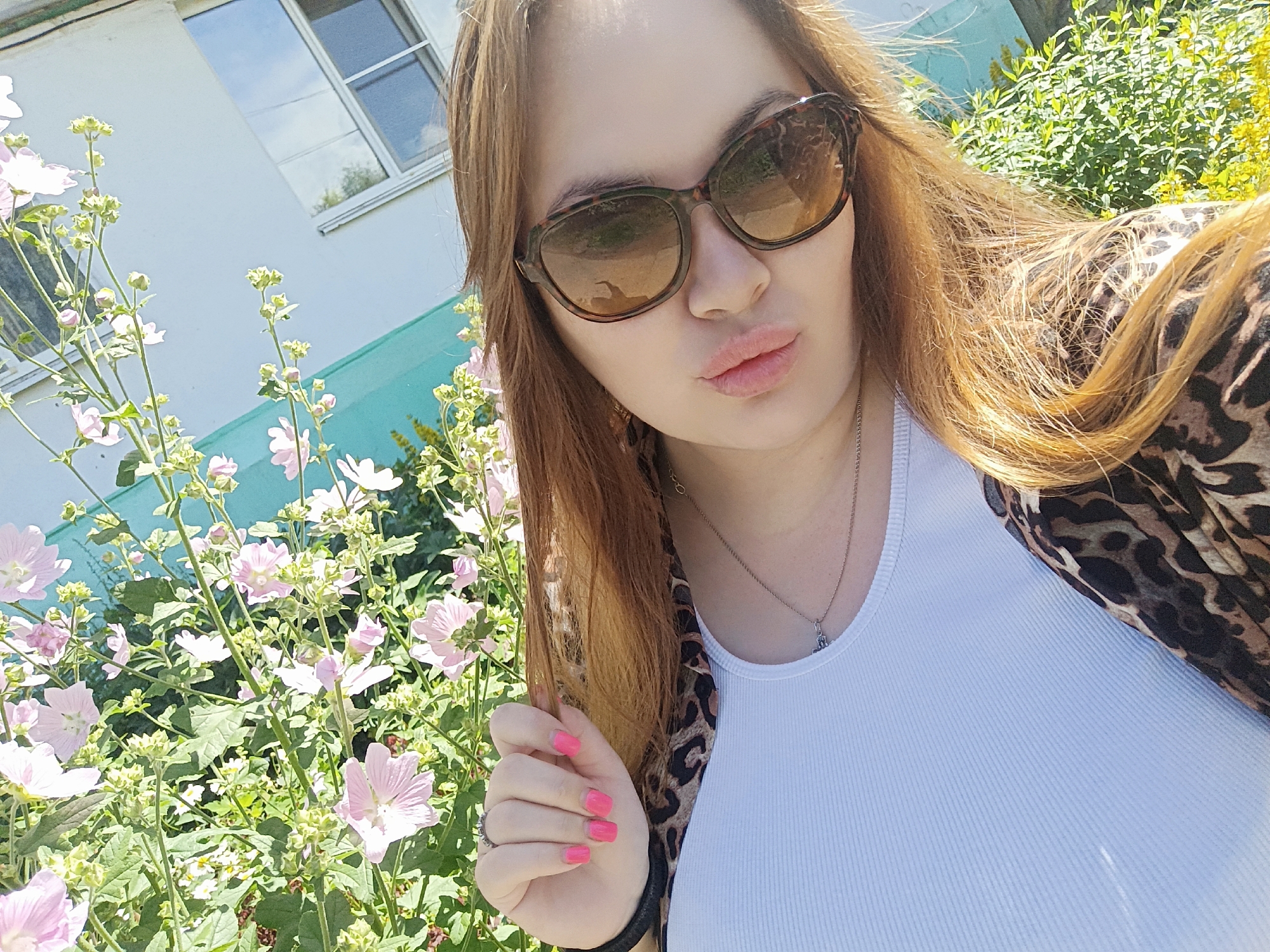 In the foreground of the image, a young woman with long, auburn hair is taking a selfie outdoors on a sunny day. She is wearing large, tortoise-shell sunglasses, a white ribbed tank top, and a leopard-print jacket. Her lips are pursed, and she is wearing bright pink nail polish. To her left, a bush with delicate pink flowers and green leaves is in bloom. In the background, a light-colored building with a window and a turquoise stripe is visible. The overall setting appears to be a residential area in the city of Chaikovsky, Russia.