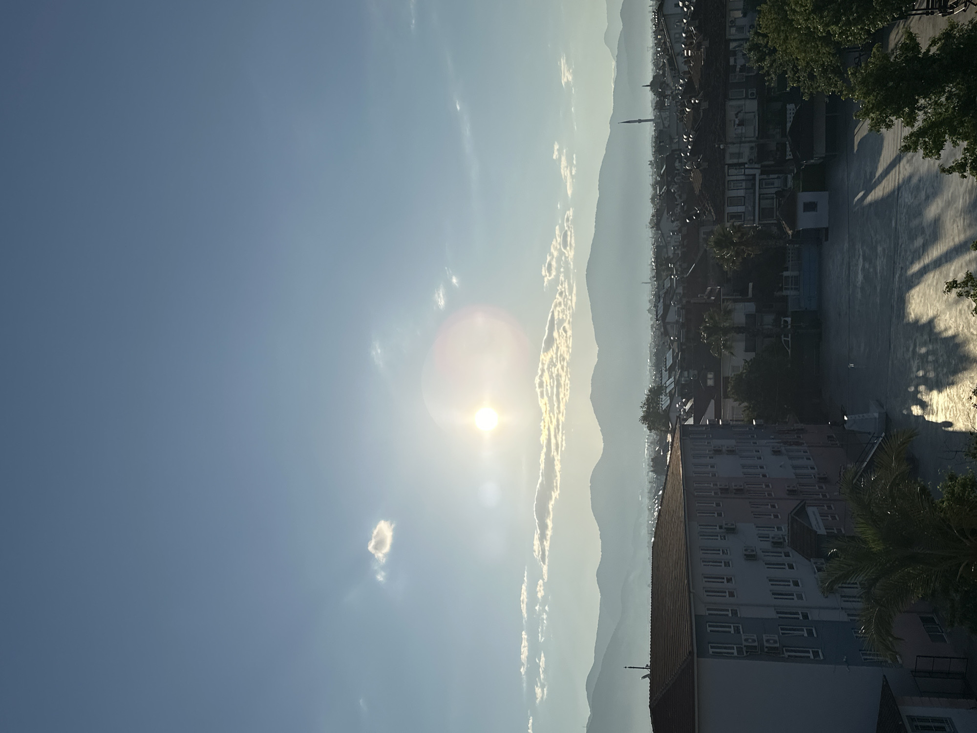 The image shows a daytime view in Fethiye, Turkey. The sky is predominantly blue, with the sun prominently visible, causing a lens flare. There are wisps of clouds present. In the distance, mountains are visible through a slight haze. The foreground contains buildings, including a taller multi-story building with many air conditioning units on its facade. Part of a courtyard or public space is also visible, with shadows suggesting trees and possibly people. A minaret is visible in the distance among the buildings. The scene appears to be shot from an elevated position.