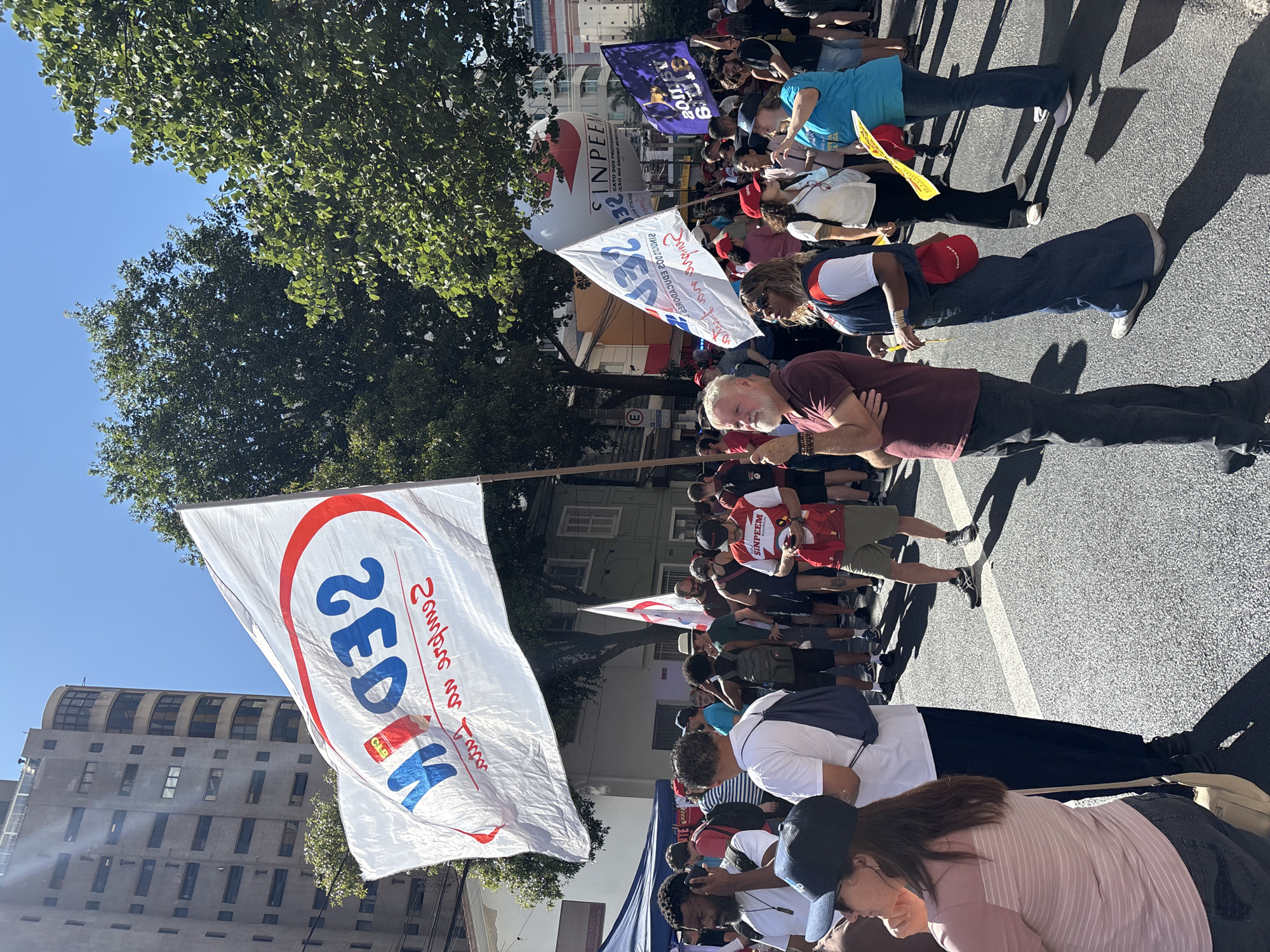 The image depicts a street demonstration or parade in the city of São Paulo, Brazil. Many people are present, holding flags and signs with political messages in Portuguese, including "ZEDEK" and "SINPEEM." The crowd appears dense, with individuals wearing a variety of clothing. A man in a maroon shirt is positioned in the foreground, looking directly at the camera. Buildings and trees are visible in the background, indicating an urban setting. The scene is brightly lit, suggesting it is daytime.