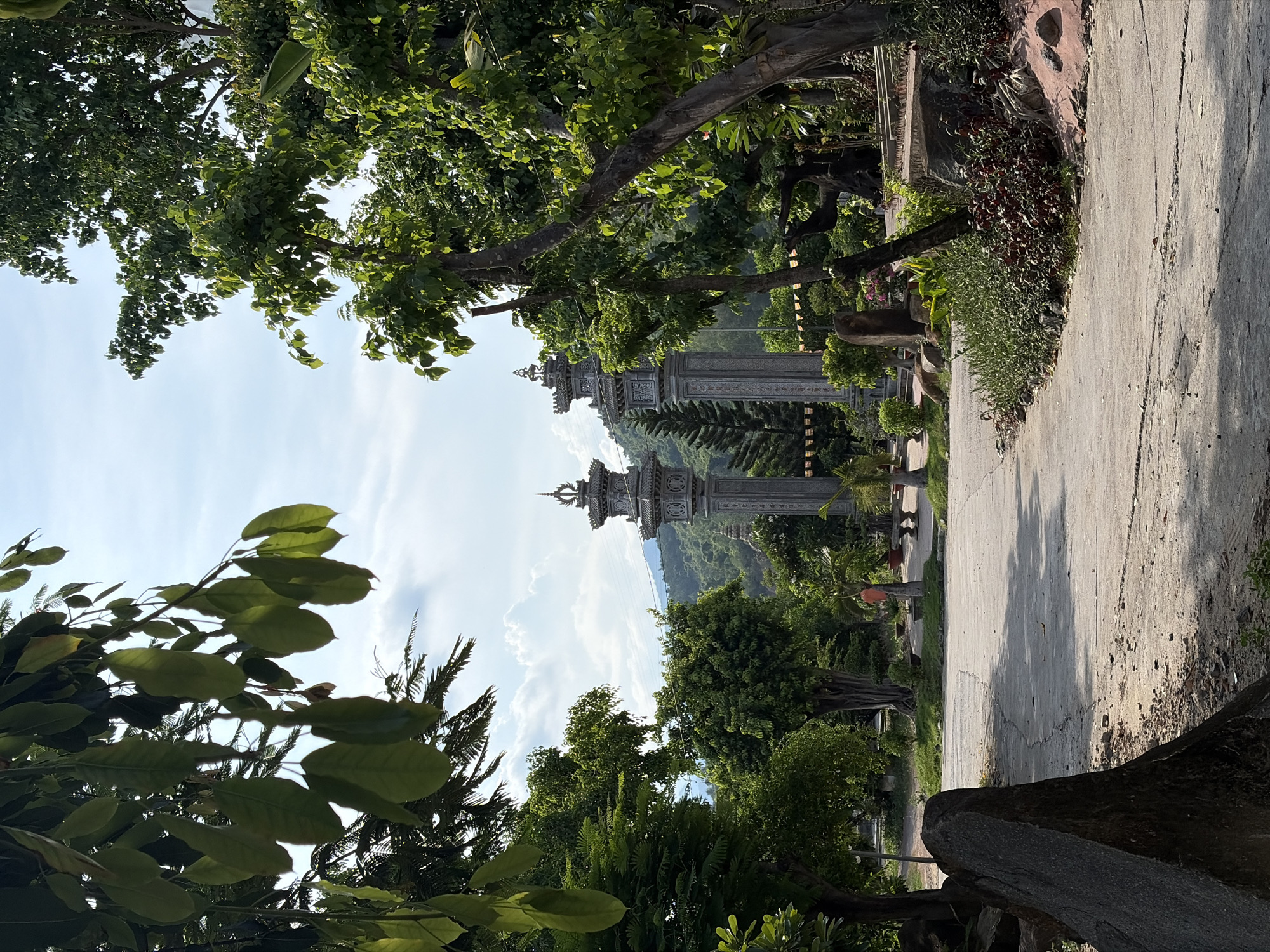 In Tay Nha Trang Ward, Vietnam, a paved pathway curves through a lush green landscape toward two ornate, stone temple gates, each topped with a tiered, decorative roof and intricate carvings. Towering trees with vibrant green leaves frame the scene on both sides, their branches reaching towards a bright, partly cloudy sky. In the distance, rolling hills covered in dense foliage rise against the horizon. The pathway itself shows signs of wear, with cracks and scattered debris, suggesting it is a well-traveled route. Shadows from the trees stretch across the path, indicating the time of day is likely late morning or early afternoon. The atmosphere is serene and natural, with the stone architecture of the temple gates providing a striking contrast to the surrounding vegetation.