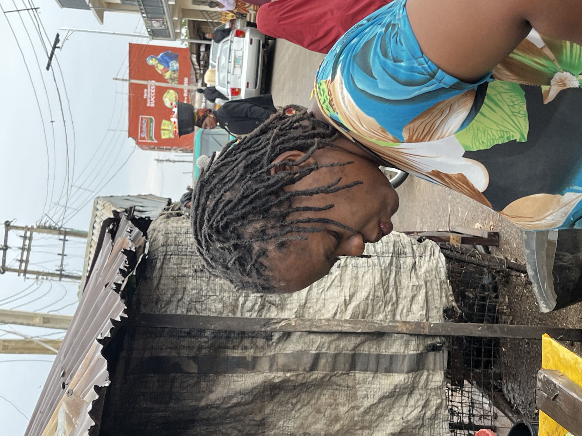 The image depicts a street scene in Jos, Nigeria. A woman with dreadlocks is captured in the foreground. She is wearing a blue floral print top. Behind her is a makeshift structure with a corrugated metal roof and a tarp-covered wall. In the background, there are other people, vehicles, and a billboard featuring an advertisement for Indomie noodles. Overhead power lines are also visible. The sky appears overcast.