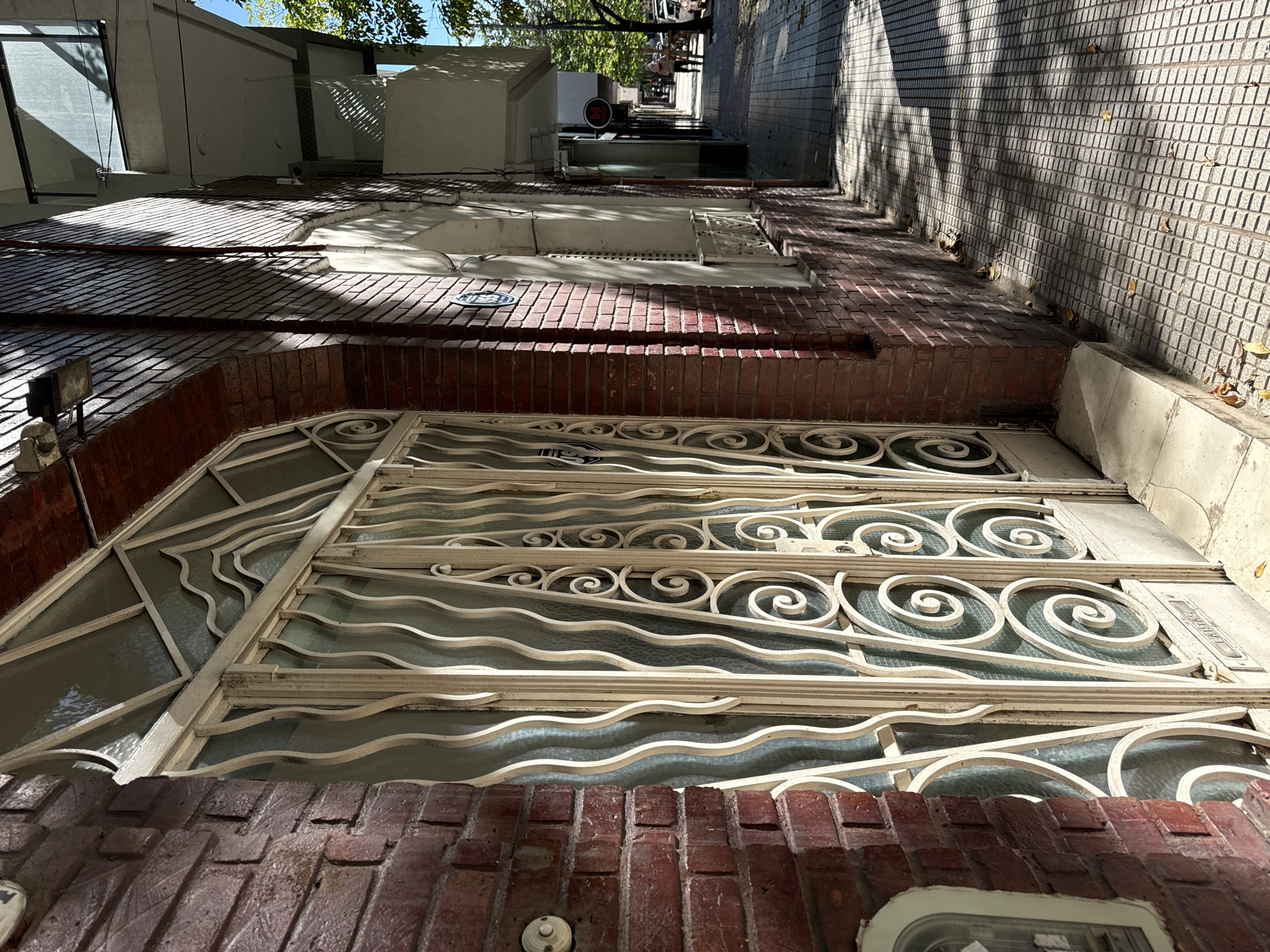 Here is a description of the image:
The image is a low-angle, street-level perspective of a building facade in what appears to be a residential or commercial area. The structure features a brick base with a large, decorative iron window covering or gate. The ironwork has an ornate, swirling pattern. Above the brick base are layers of the building, including additional windows and architectural details, extending upwards and receding into the distance. The perspective creates a vanishing point towards the end of the street.
The right side of the image shows a textured wall with shadows cast by trees. The overall light suggests a sunny day. Given the architectural style and general appearance of the image, it is very likely located in the city of Buenos Aires, Argentina.
