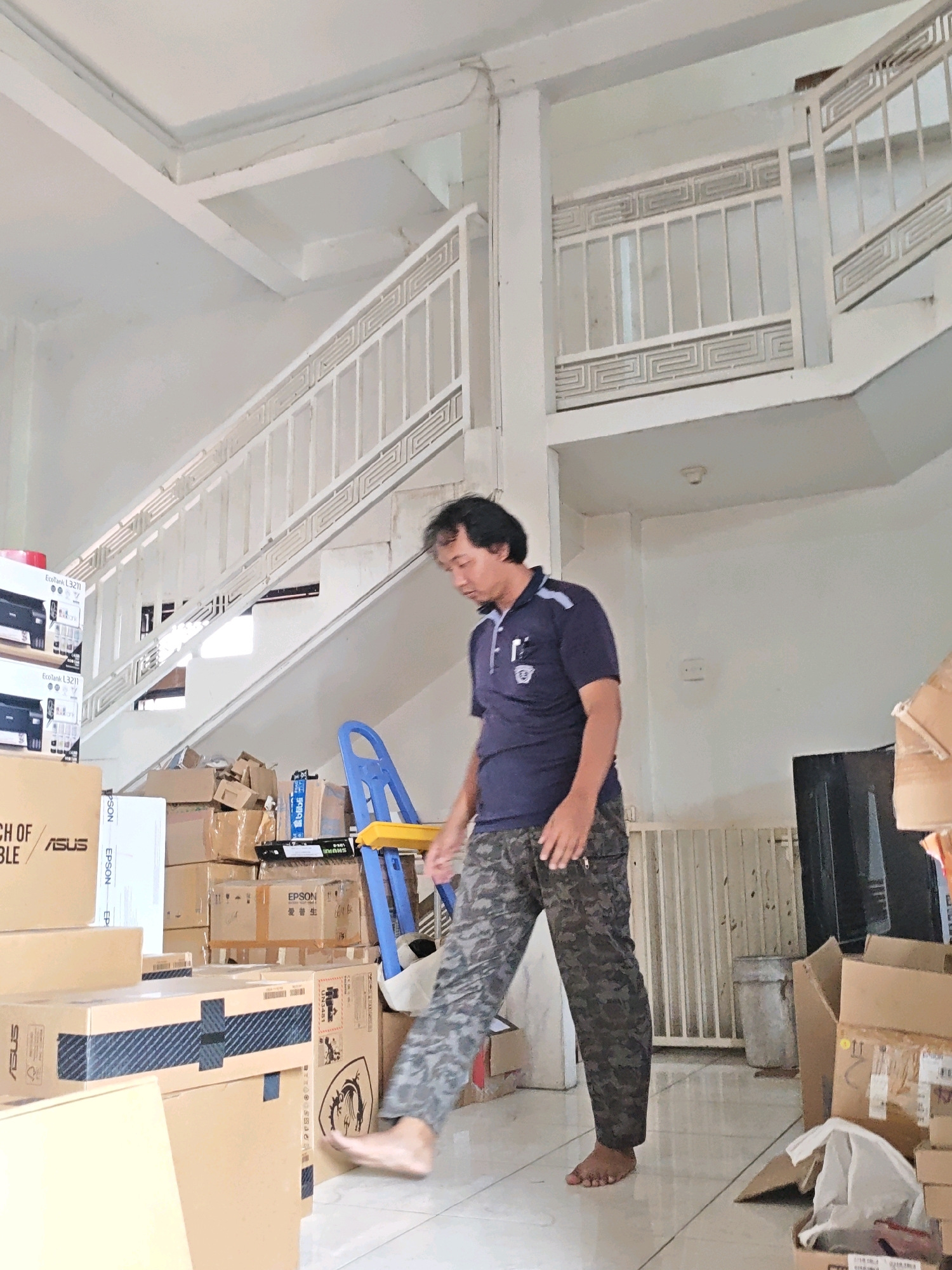 The image shows an indoor scene, likely a warehouse or storage area, in Ngasem, Indonesia. A middle-aged man wearing a dark blue polo shirt and camouflage pants is walking. He is barefoot. The environment is filled with cardboard boxes of various sizes, some stacked high. In the background, a white staircase with a decorative railing is visible, extending upwards. There is also a blue and yellow step stool or ladder. The lighting suggests it is daytime.