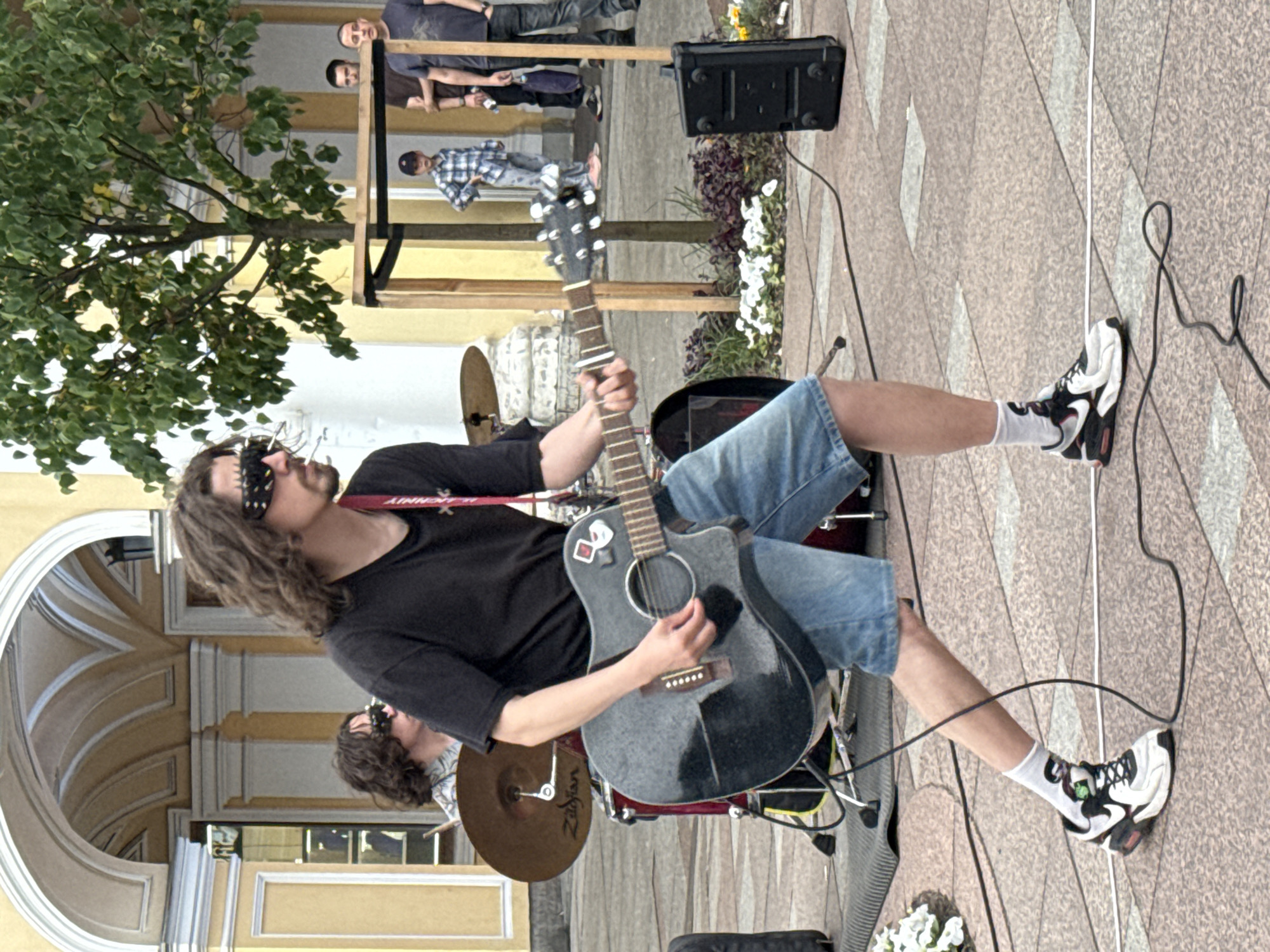 The image depicts an outdoor street performance in Saint Petersburg, Russia. In the foreground, a young man with long, wavy brown hair, wearing a black t-shirt, denim shorts, white socks with graphics, and black and white athletic shoes, is playing an acoustic guitar. He has on distinctive studded sunglasses and a red strap is visible around his neck. Behind him, another individual with similar hair and sunglasses is seated behind a drum set, indicated by cymbals and drum heads. The background features architectural elements characteristic of Saint Petersburg, including arched windows and yellow-toned buildings. Several onlookers are also present, including a child in a plaid shirt and pants standing near a wooden structure, and two men standing further back. The ground is paved with cobblestones, and some greenery and flowers are visible in the scene. A black speaker sits on the ground to the right.