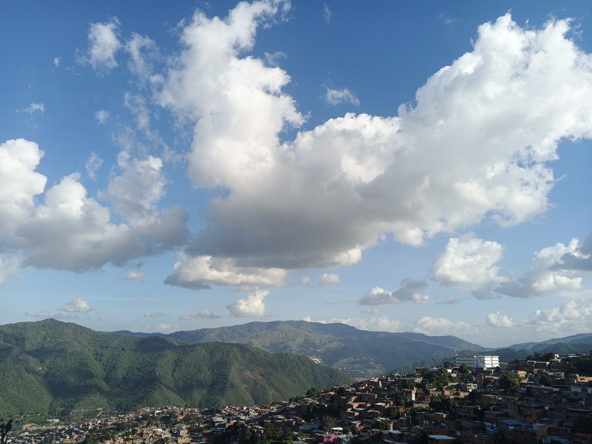 The image shows a panoramic view of Caracas, Venezuela, on a sunny day. A vibrant blue sky is dotted with white, fluffy clouds that dominate the upper half of the scene. Below, a dense urban landscape spreads across the hilly terrain. Many densely packed red-tiled roofs of buildings stretch into the distance, marking the cityscape. Green, rugged mountains form the backdrop, providing a natural contrast to the urban development. A large, white building complex stands out amidst the residential area. No people are visible in the image.