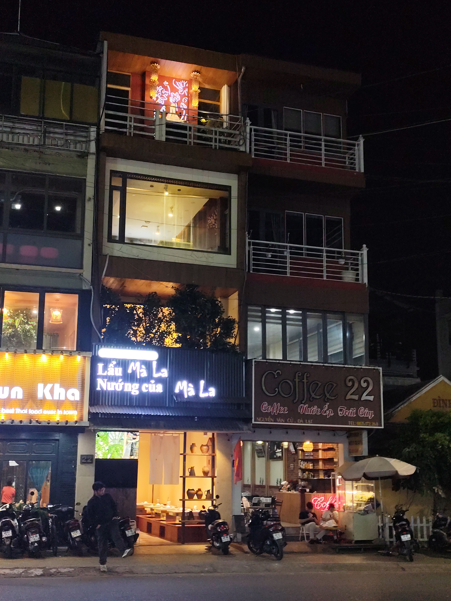 The image shows a street scene at night in Da Lat, Vietnam. The scene is dominated by three adjacent storefronts: "un Kha" (Thai food), "Lẩu Mà La - Nướng của Mà La," and "Coffee 22." Several motorbikes are parked along the curb in front of the businesses. A person wearing dark clothing is walking across the street near the "Lẩu Mà La" restaurant. People are sitting outside the coffee shop underneath an umbrella. The buildings are multi-storied with balconies. The street name "NGUYỄN VĂN CỪ" is included in the text.