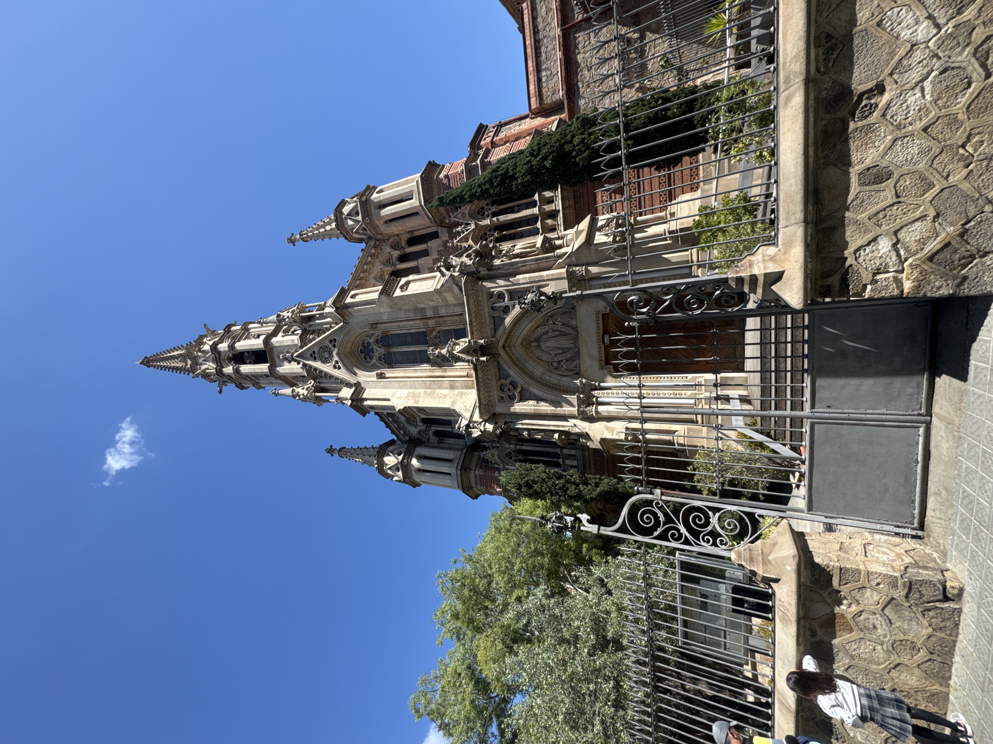 The image depicts a building with ornate Gothic architecture in Barcelona, Spain. The building is tall and slender with a pointed spire. The exterior is light-colored stone with detailed carvings and windows. A wrought iron fence runs along the front of the building, with an open gate on the right side. The fence is partially obscured by green trees and foliage. The sky is bright blue with a few white clouds. A section of stone patterned fence wall is along the lower right side of the image. The bottom right of the image shows a person wearing a white and gray shirt with black leggings.