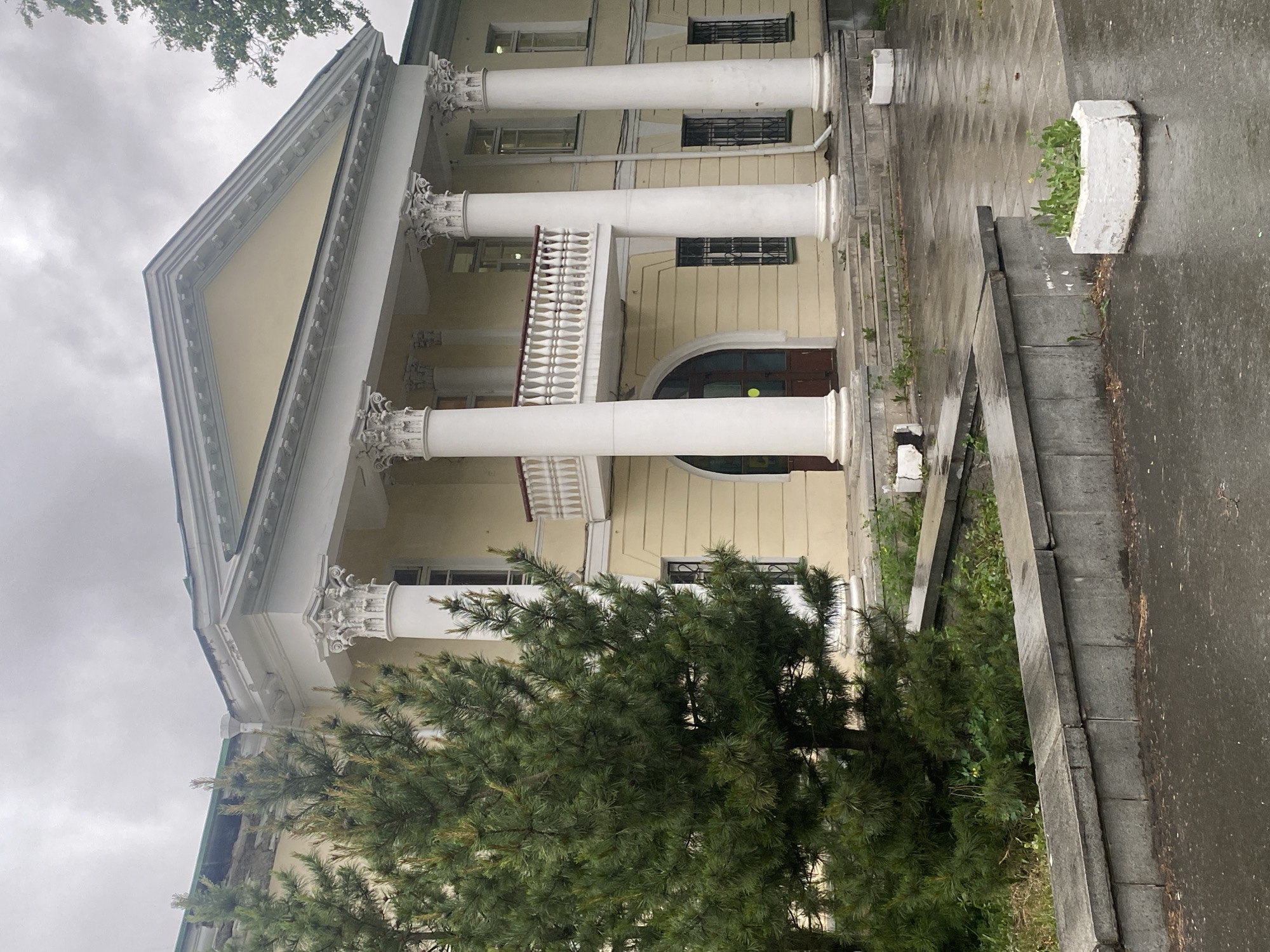 The image shows the facade of a building with classical architectural elements. The building is light yellow with white columns, a balcony, and a triangular pediment. Windows are visible on multiple floors. In the foreground is a green coniferous tree partially obscuring the building's lower section. A set of steps leads up to the building's entrance, with large concrete planters positioned along the steps. The ground appears wet, possibly from rain. The sky is overcast.
Based on the architectural style and the available information, it is likely the location is in Yekaterinburg, Russia, but the exact building name or address cannot be determined from the image alone. There are no people visible in the image. The primary objects are the building, columns, tree, steps, planters, and sky. The action is static, showing a scene with no immediate activity. The environment is characterized by classical architecture and potentially rainy weather.