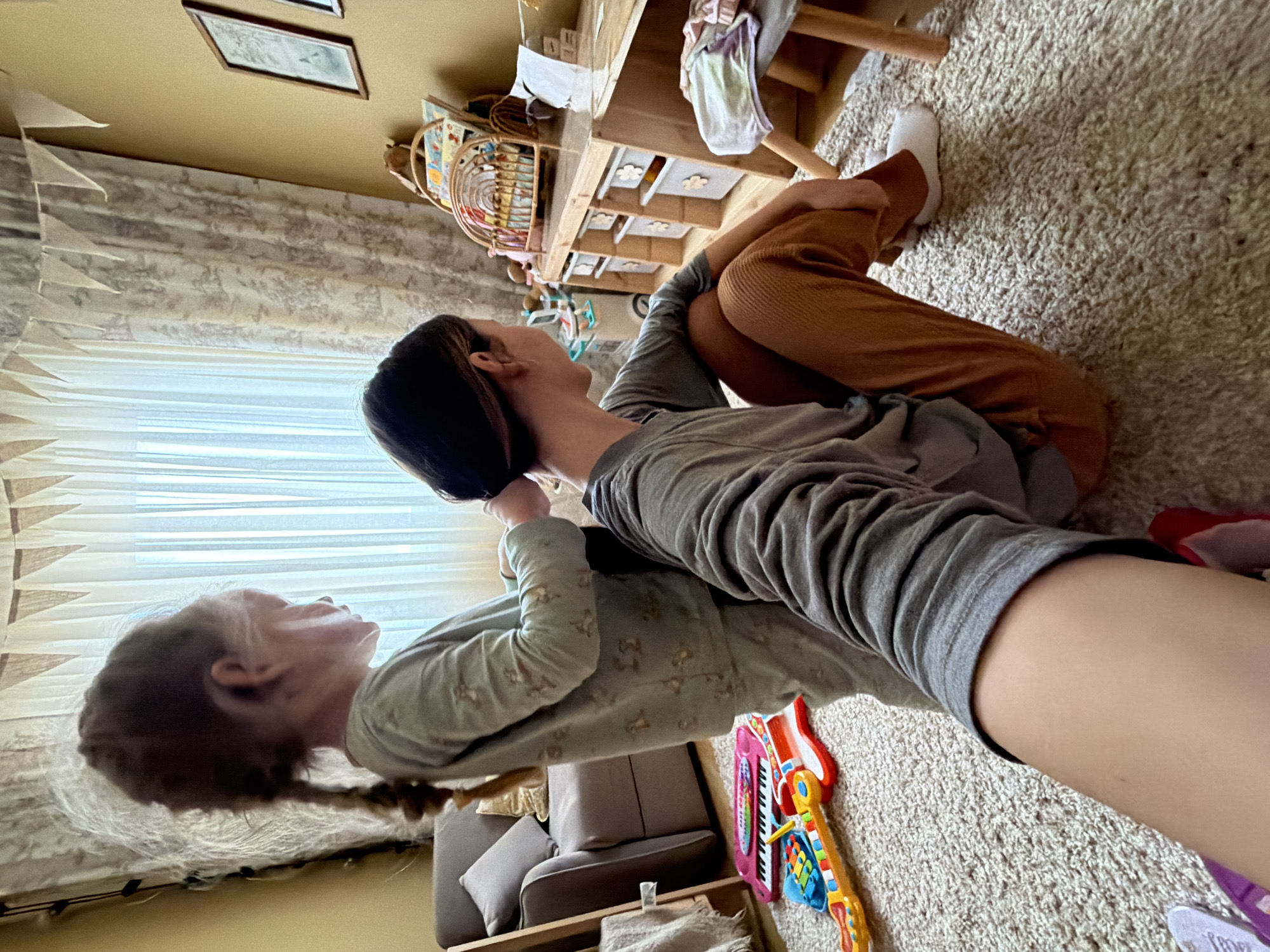 Two individuals are lying on a beige carpet indoors, one atop the other. The lower individual is fair-skinned with light brown hair in a braid, wearing light green pajamas with a pattern. The upper individual has dark hair and wears a gray, long-sleeved shirt and brown pants. A white sock is visible on one foot. A wooden toy chest with drawers and a wicker basket filled with items are near the pair. A toy guitar and keyboard are near the pair. The room has off-white walls, a window with sheer curtains, and pictures on the wall.
There is no indication this photo was taken in Khimki, Russia.