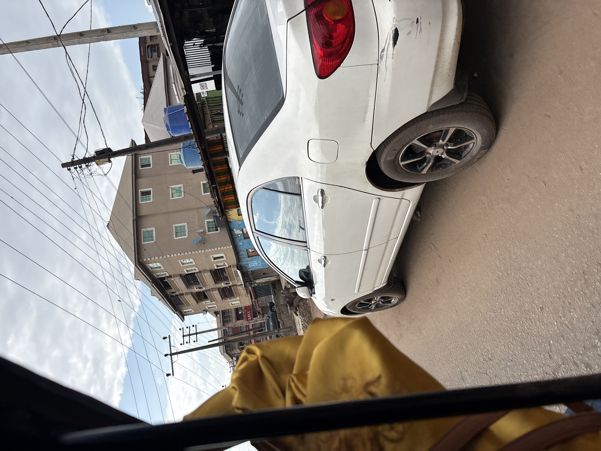 The image depicts a street scene in Nkpor, Nigeria, viewed from the perspective of someone within a vehicle, possibly a rickshaw or tuk-tuk, indicated by the presence of a yellow fabric and dark framing in the foreground. A white sedan, likely a Toyota Corolla based on its taillight design, is the prominent subject, positioned on a dirt and asphalt road. The car shows visible signs of wear, including dents and scratches on its rear bumper and body panels. Behind the car, several multi-story buildings are visible, characterized by their beige and brown facades, tiled roofs, and numerous windows, some with green shutters. Blue water tanks are mounted on some rooftops. Overhead, a complex network of electrical wires and utility poles crisscrosses the sky. The environment appears to be an urban setting with a mix of residential and commercial structures. The lighting suggests it is daytime, with a partly cloudy sky. In the background, another dark-colored car is partially visible on the road.