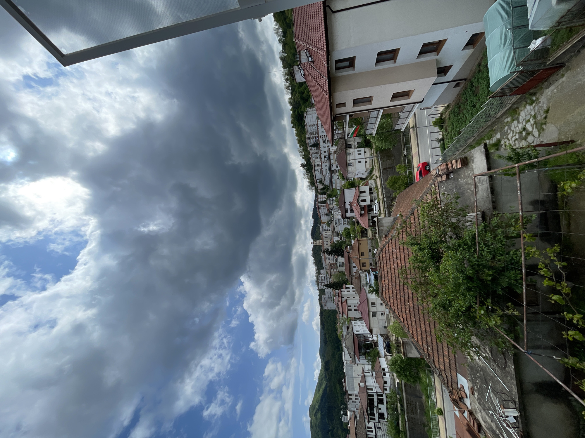 An eye-level, vertical oriented shot presents a residential area in Zlatograd, Bulgaria. In the background, numerous white and beige houses with red tiled roofs are nestled on a sloping hill, backed by a dense forest. The sky is predominantly covered with heavy, grey clouds, with patches of blue sky and white clouds scattered in between. Foreground details include sections of buildings, a metal fence, and greenery, indicating a view from an elevated position, possibly a balcony. No people are visible in the image.