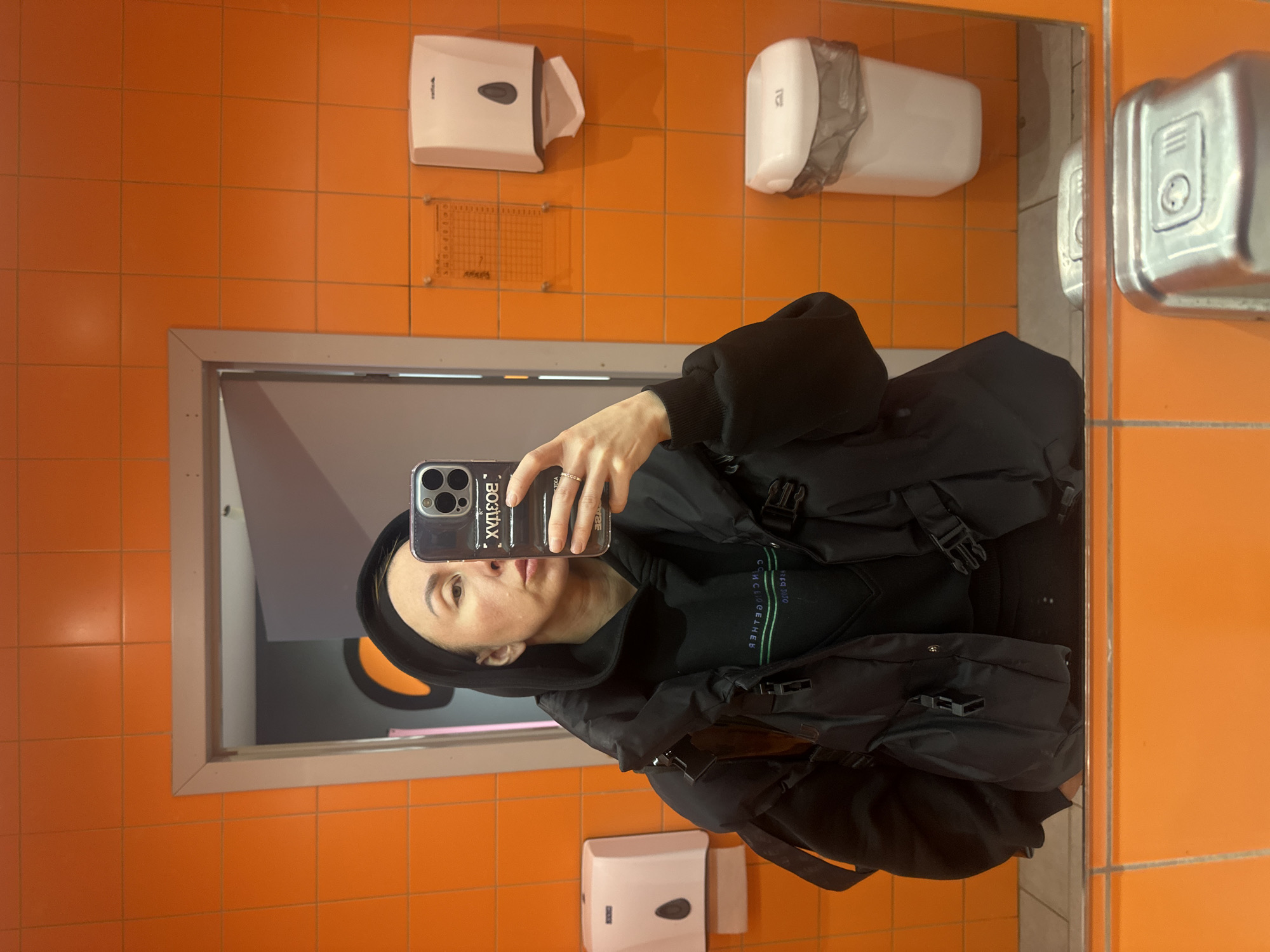 A person is taking a mirror selfie in a bathroom with orange tiled walls. The bathroom contains a paper towel dispenser, a hand dryer, and an additional dispenser of some kind. The person is wearing a black hoodie and jacket, and appears to have a backpack on. The location and city of Votkinsk, Russia are not discernable from the image.