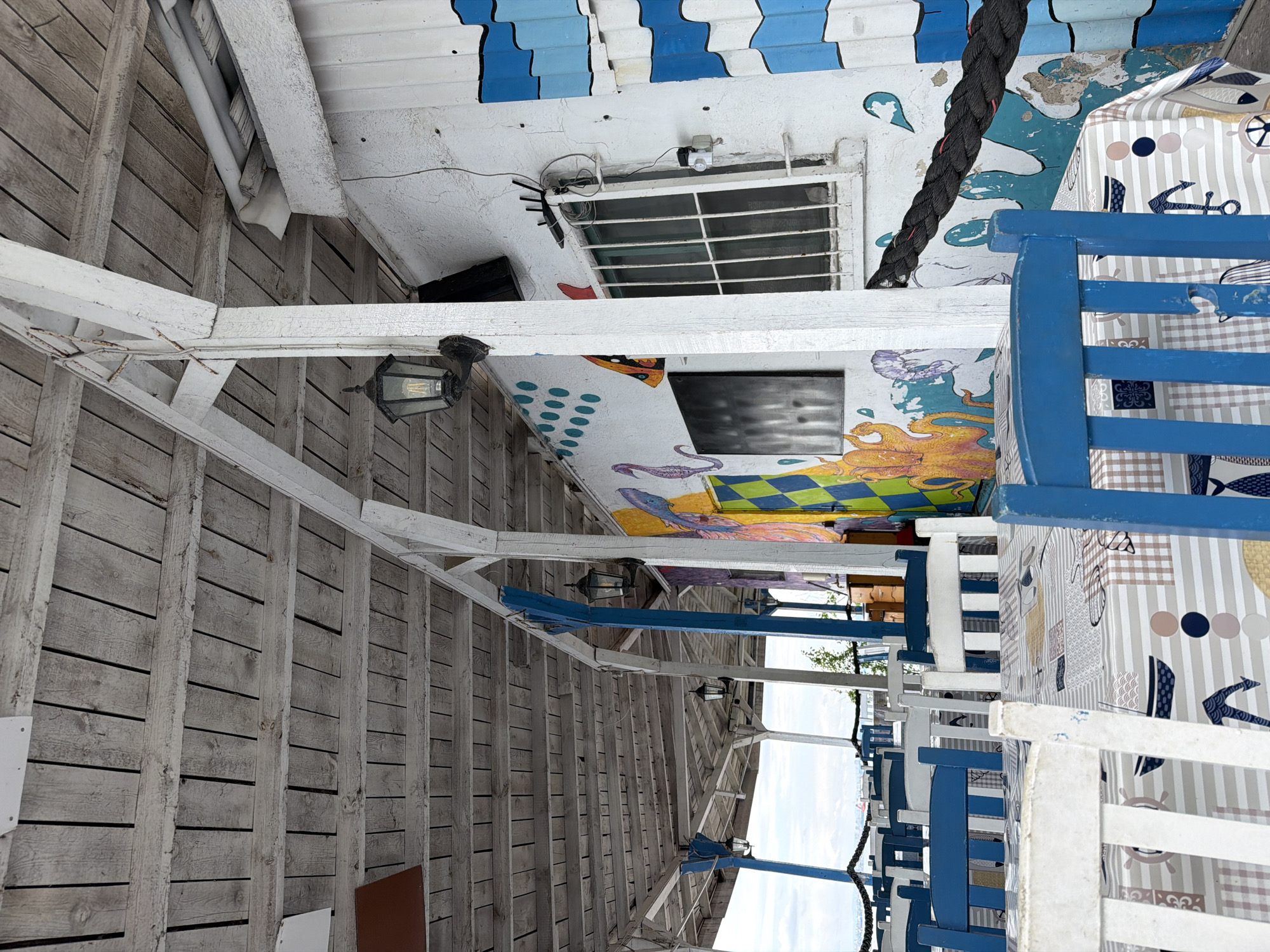 The image shows the interior of an outdoor restaurant in Burgas, Bulgaria. The structure has a wooden framework and ceiling. The ceiling is decorated with marine-themed paintings and blue and white patterns. A security camera is visible on the ceiling. Tables are set with tablecloths featuring nautical motifs. Blue and white chairs surround the tables. A black rope is present. The view outside suggests the restaurant is near the sea. The lighting indicates it is daytime.