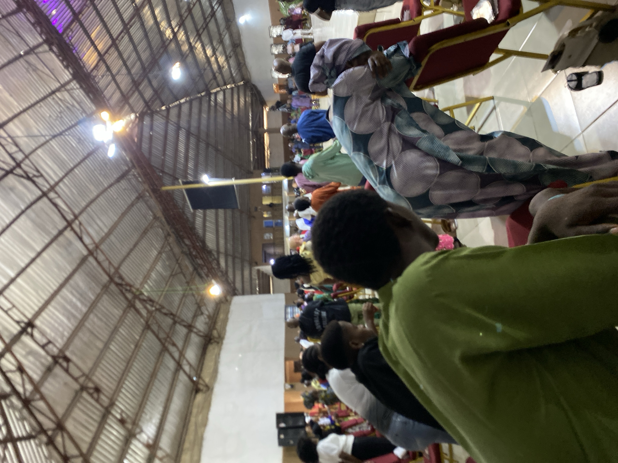 The image shows an interior space, potentially a church or large hall, with rows of people seated in chairs. The perspective is tilted, giving an upward view of the ceiling structure, which appears to be made of metal sheeting and supporting beams with lighting fixtures attached.
People are of varying skin tones and wearing different colored clothing. Some individuals are in the foreground, with a person in a green shirt being most prominent. Further down the rows, people are wearing blue, white, black, and patterned clothing. Most are seated and facing the same direction.
Chairs are arranged in rows, with red seats and metal frames. The floor is light-colored.
The location is tagged as Unknown, Unknown.