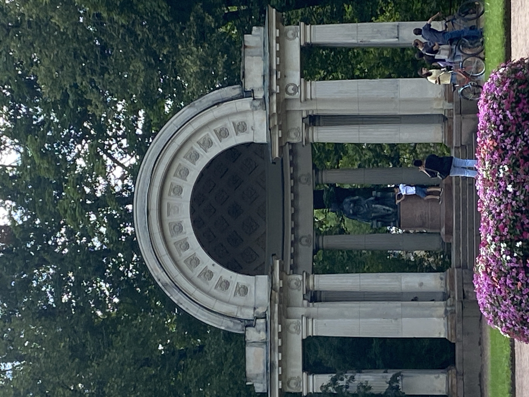 In the city of Pavlovsk, Russia, a monument featuring a rotunda with classical columns and an arched roof stands amidst lush green trees. The rotunda houses a bronze statue of a seated woman. In front of the monument, a large, vibrant flower bed bursts with purple and pink blossoms. Several people are present: one person is climbing the steps of the monument, another is taking a photograph, and a group of four is gathered with bicycles. The scene appears to be a public park or garden, with a lawn and paved walkway visible.