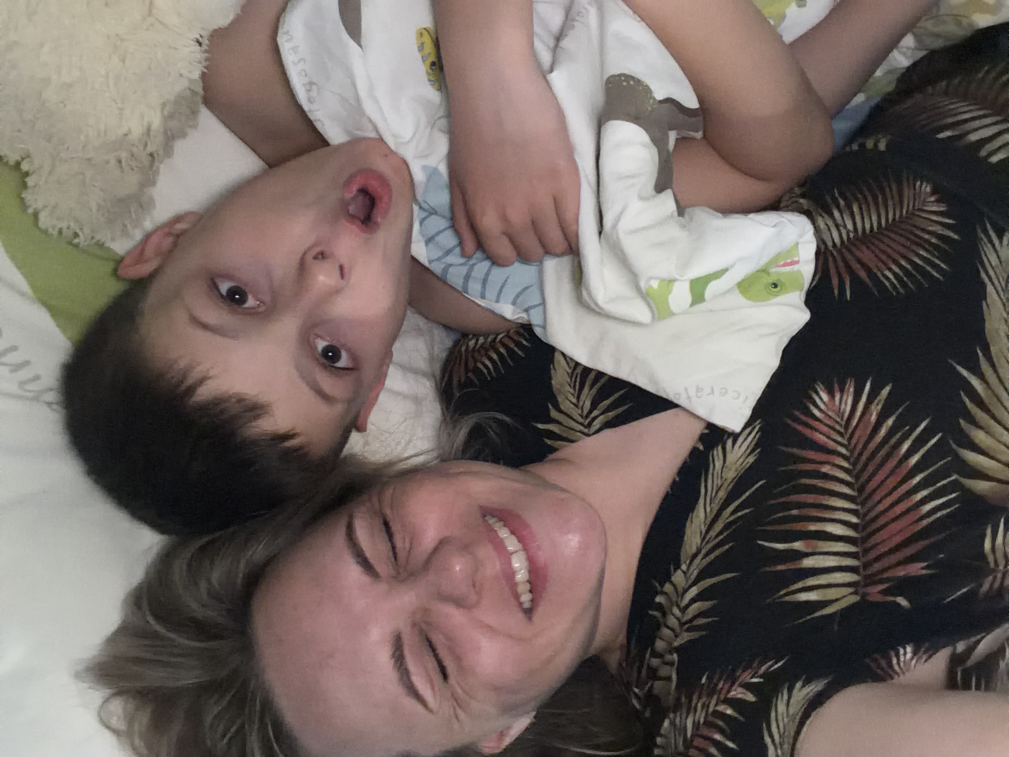 A woman and a child are lying down in an indoor setting. The woman is smiling with her eyes squinted shut. She wears a dark-colored top with a pattern of palm leaves in shades of gold and brown. The child, who appears to be male, is positioned above the woman, with his head near her shoulder. He has his mouth open in an apparent expression of surprise or playfulness. The child is wearing a white top with cartoon-like images. Part of a white plush toy is also visible near the child. The background suggests they are on a bed with white and green patterned bedsheets. The location in the city of Votkinsk, Russia is not discernible from the image.