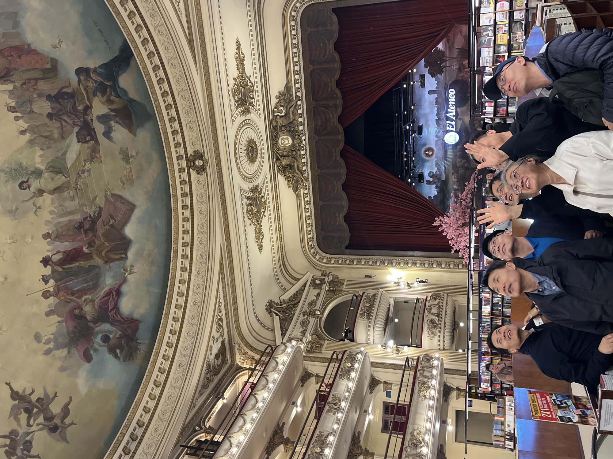The image depicts El Ateneo Grand Splendid, a bookstore in Buenos Aires, Argentina, located in a converted theatre. The image is oriented to highlight the architectural details of the space. On the left, a mural-covered ceiling is visible, featuring classical figures and ornate borders. The theatre's balconies and decorative plasterwork are visible. Red curtains frame the stage, which now serves as a reading area with bookshelves and displays. A group of people are standing on the right, oriented to face the camera.