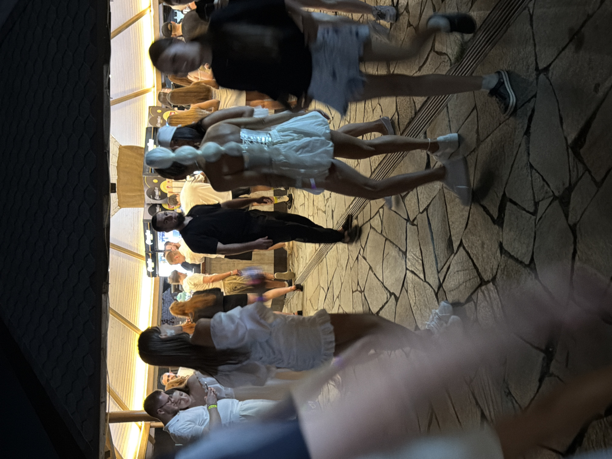 The image depicts a nighttime scene with a group of people in what appears to be an outdoor venue, possibly a bar or club, with a covered area. The ground is paved with irregular stone tiles. In the foreground, a person's arm and shoulder are prominently blurred, suggesting the photographer is also present and close to the scene.
Further into the image, several individuals are visible. On the left, a man with glasses and a white shirt is looking at his phone, accompanied by a woman. Next to them, a woman in a white off-the-shoulder dress with ruffled trim is facing away from the camera.
Towards the center, a person with a distinctive white hairstyle is wearing a white corset-style top and a short skirt. They are standing on the stone pavement.
To the right, a woman in a black top and a light-colored skirt is walking away from the camera. Other individuals are present in the background, some near what looks like a bar area with glowing lights. The overall atmosphere suggests a social gathering or event. There is no specific information within the image to indicate the location is the city of Engels, Russia.