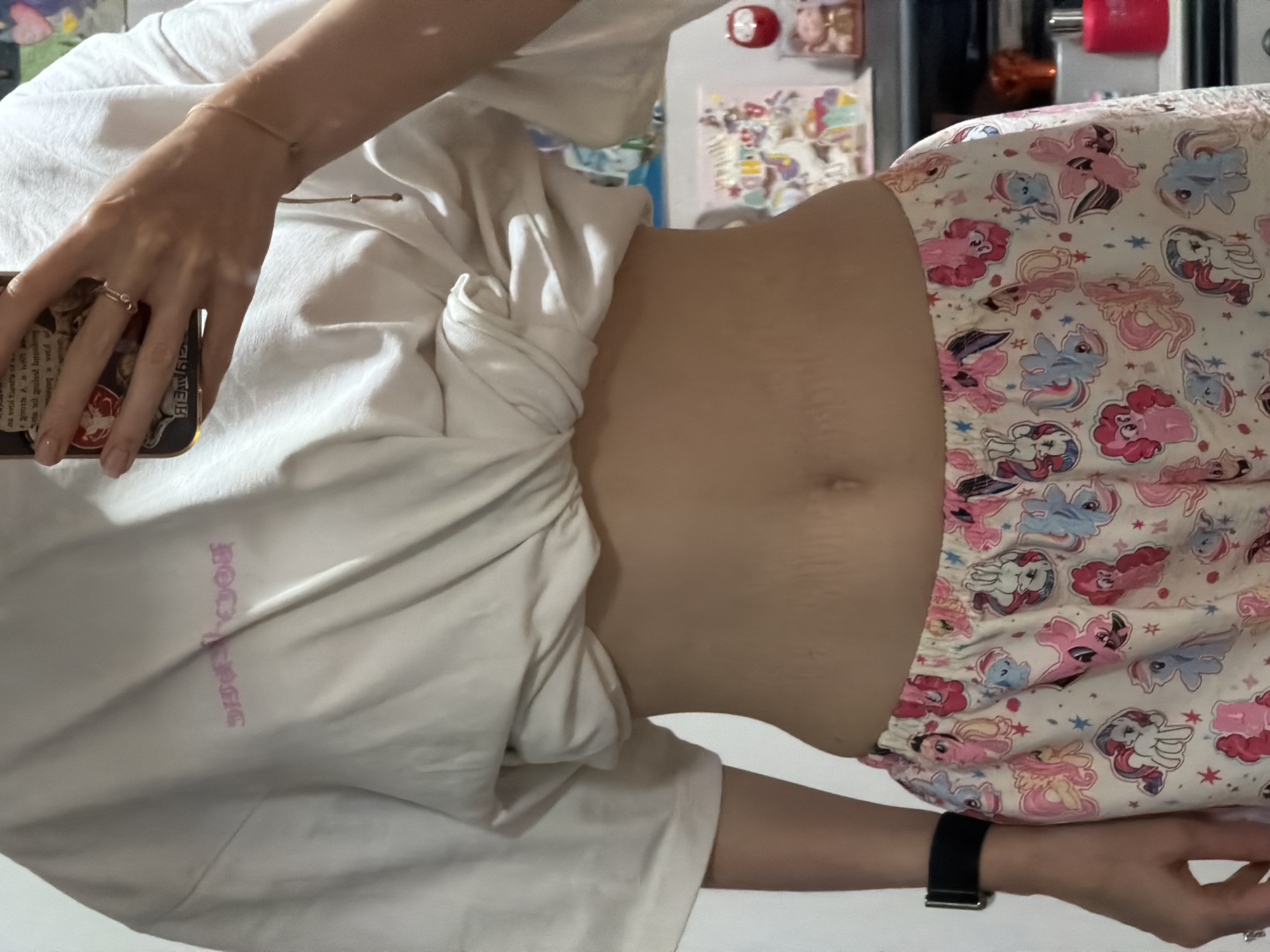 A person's midsection is visible in this medium shot image. They are wearing a white t-shirt tied at the waist and patterned pajama bottoms featuring cartoon ponies. The person has stretch marks on their stomach. They are also wearing a black smartwatch on their left wrist and a ring on their right hand. The background is out of focus but appears to be an indoor setting with shelves of items, including decorative figures. The image does not contain any information about the location in the city of Moscow, Russia.