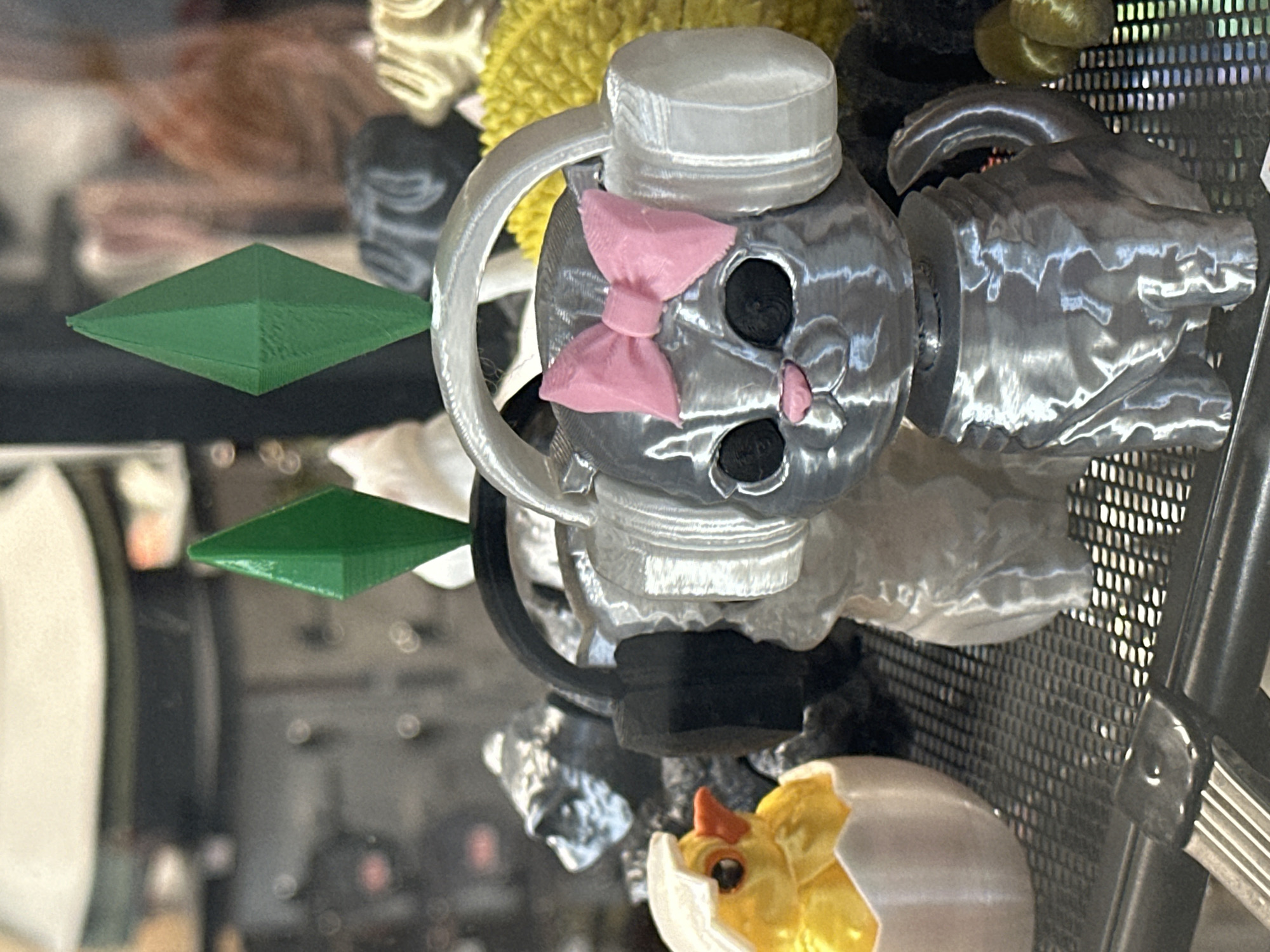 The image shows a collection of 3D-printed objects displayed inside a glass cabinet. The focal point is a silver, metallic-looking cat-shaped figurine with a pink bow on its head and a bottle cap-like structure on top. Other visible objects include a green diamond-shaped object, a yellow fluffy sphere, a silver figure resembling a hand or dragon head, and a chick hatching from an egg. The objects are arranged on multiple levels within the cabinet. The background is blurred due to the depth of field, but appears to be a room or building. The description does not provide any evidence of a location in the city of Novosibirsk, Russia.