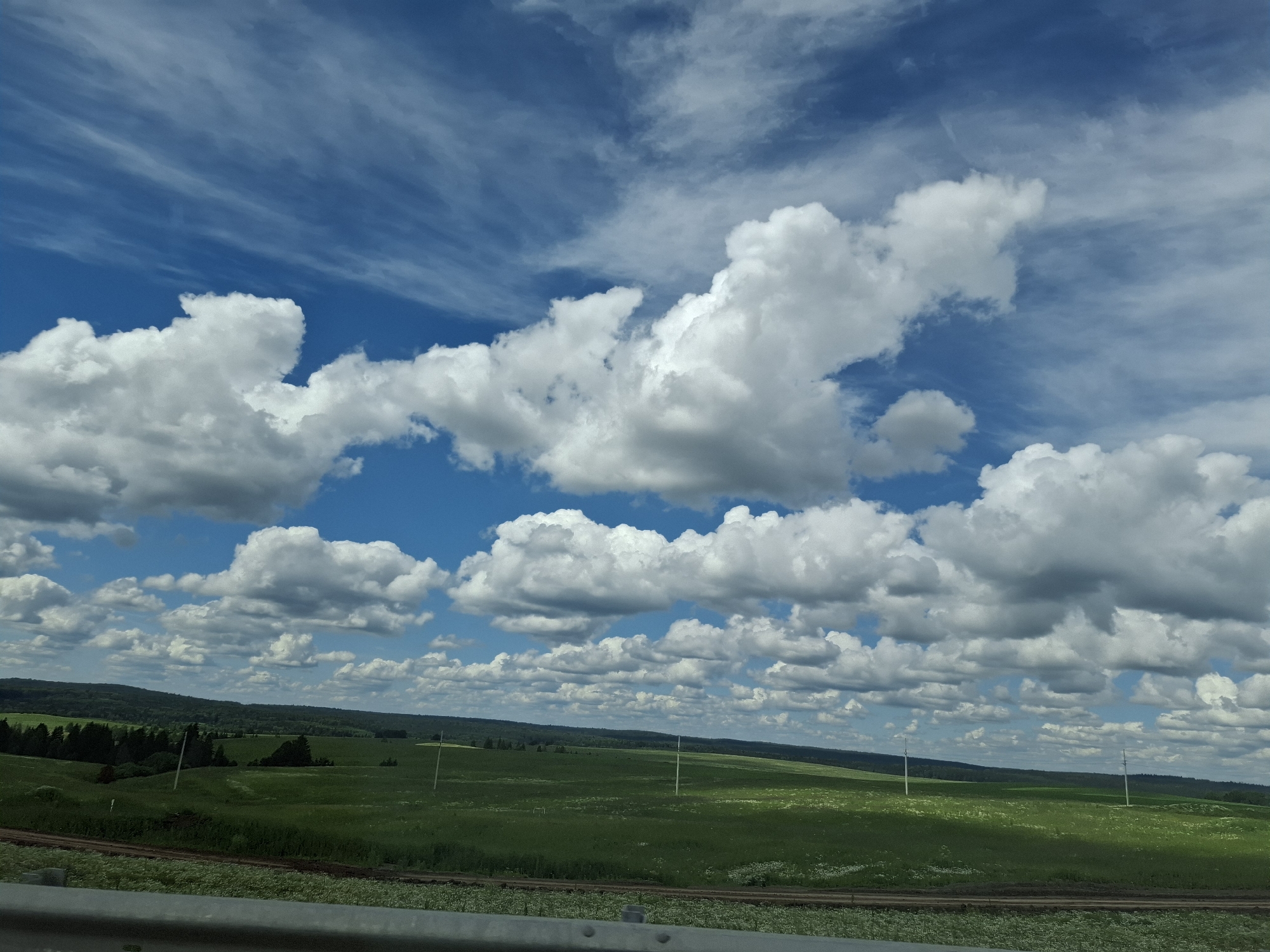 The image depicts a vast, rolling green landscape under a dynamic blue sky filled with white, puffy cumulus clouds and wispy cirrus clouds. The foreground shows a weathered metal guardrail. Beyond the guardrail, a dirt path runs horizontally across the scene, bordered by low-lying vegetation. The middle ground consists of expansive green fields, possibly farmland or pasture, dotted with occasional white wildflowers. Several utility poles are visible in the distance, rising from the fields. The background features a dense line of dark green trees forming a treeline against rolling hills, which are shrouded in a light haze. The overall scene suggests a rural, possibly agricultural area in or near the city of Уразгильды, Russia, on a bright, partly cloudy day. There are no people or animals visible in the image.