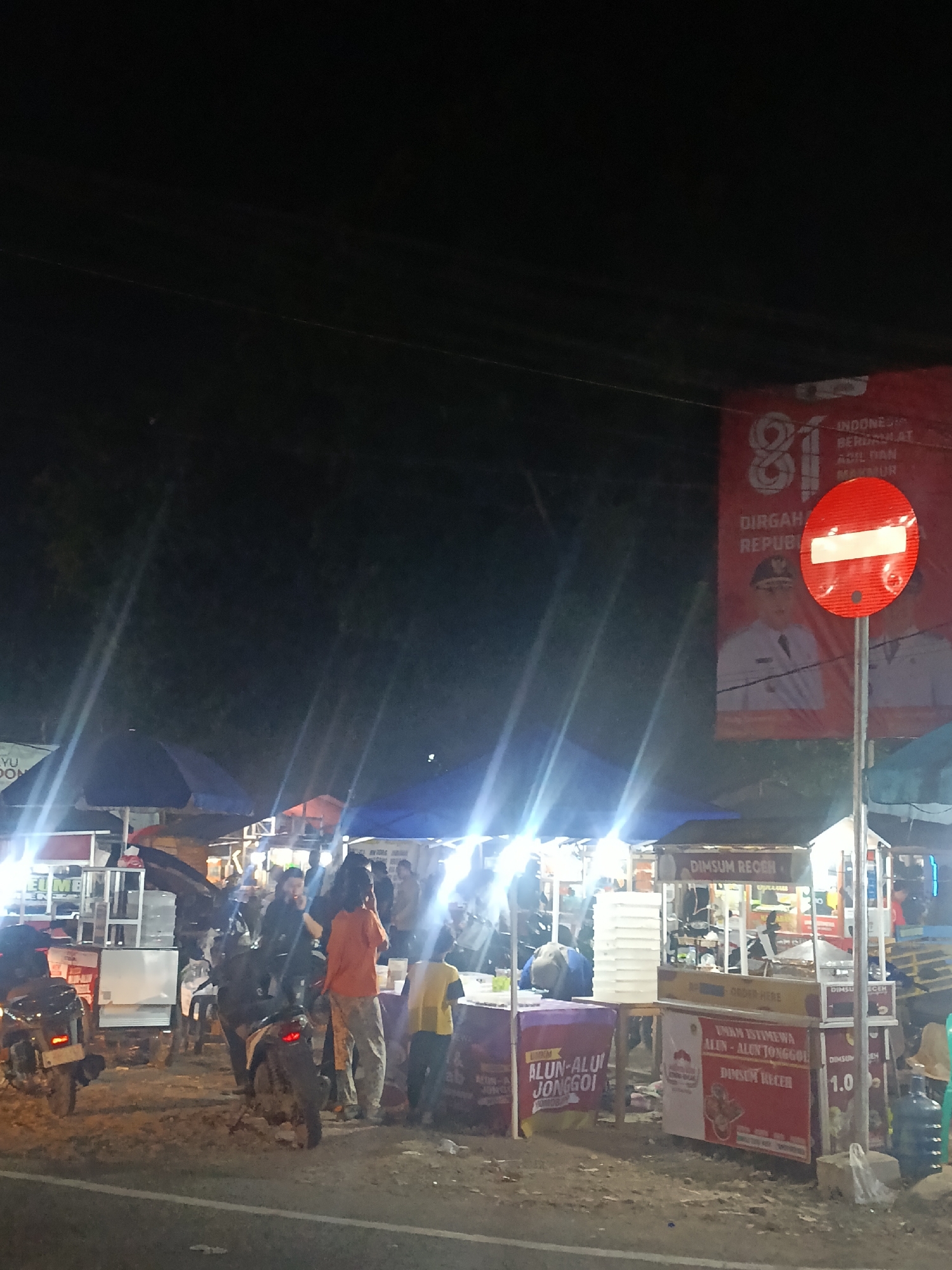 At night in Jonggol, Indonesia, a street food scene unfolds under the glow of artificial lights. Several food stalls are set up, their awnings illuminated, creating a lively atmosphere. One stall prominently displays the words "DIMSUM RECEM" and "ALUN-ALUN JONGGOL." Another stall has signage that says "UMM ISTIMEWA ALUN-ALUN JONGGOL DIMSUM RECEM." A large red and white sign with the number "81" and an image of a person in official attire is visible in the background, likely related to a national celebration. A red circular traffic sign with a white horizontal bar indicates "no entry." Several motorcycles are parked near the stalls, suggesting this is a popular gathering spot. People are seen interacting at the stalls, with some standing and others milling about. The ground appears to be a rough, unpaved surface. The overall impression is a vibrant, bustling night market in Jonggol.