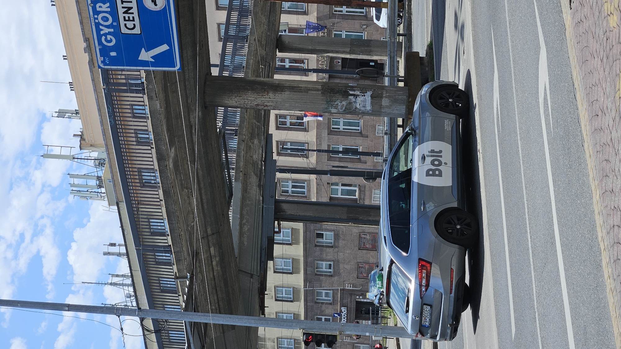 The image shows a grey Skoda Superb estate car with "Bolt" branding on its side, driving on a street in Bratislava, Slovakia. The car has a license plate that reads "PN 900EH". The car is in the foreground, and a large concrete overpass structure is visible behind it. Several buildings with many windows are also in the background. There are traffic lights on the left side of the image. A blue directional sign indicating "GYOR" and "CENT" with an arrow pointing to the right is also visible. The sky is blue with scattered clouds.