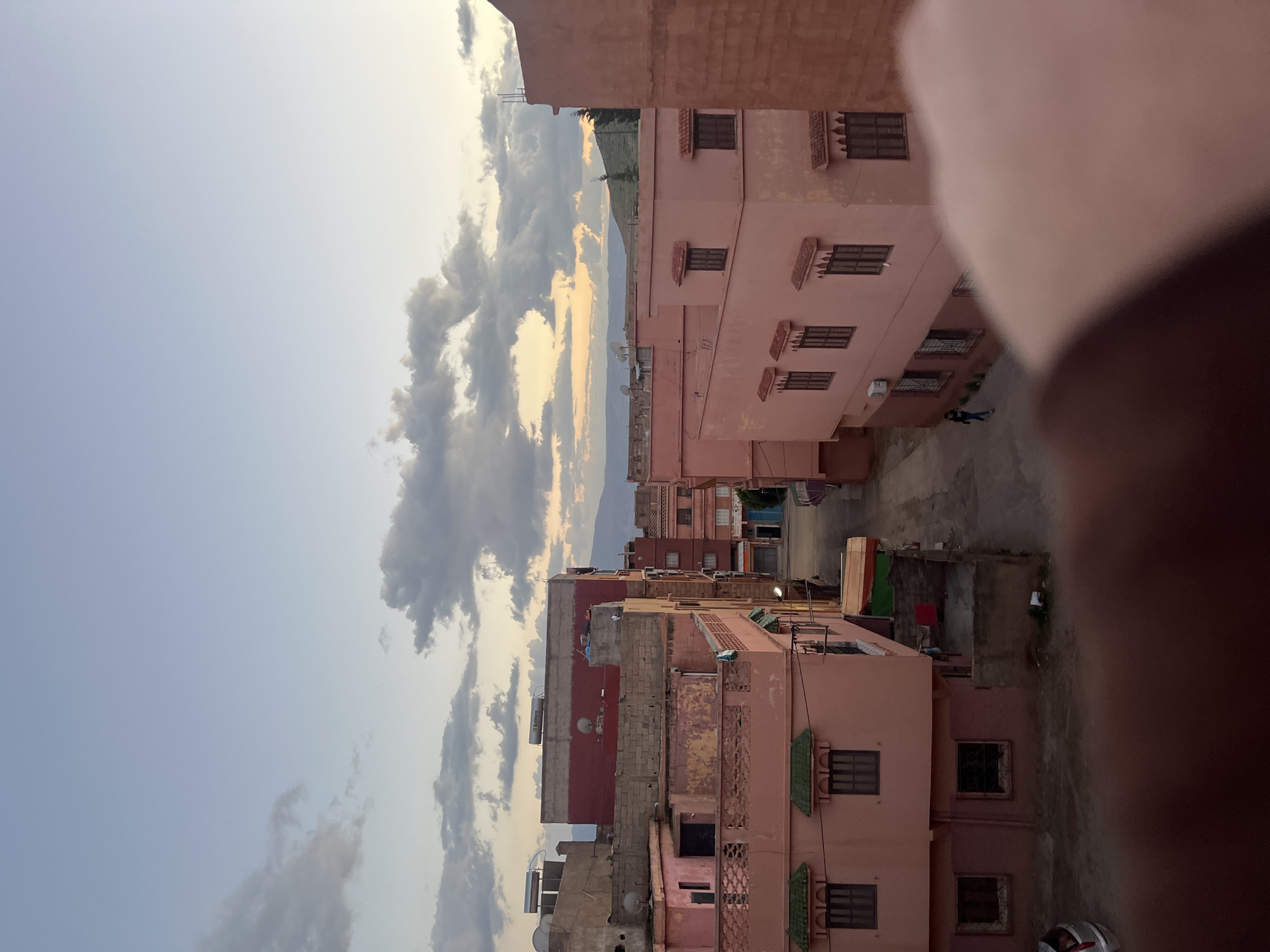The image is taken from an elevated position in the city of Ouaouizert, Morocco, looking down on a street lined with pink buildings of varying heights and architectural styles. The buildings have windows with dark shutters, satellite dishes, and water tanks on their roofs. A figure is walking on the street. The sky is filled with clouds, with a glimpse of mountains in the distance. Part of the photographer's hand is in the frame and out of focus.