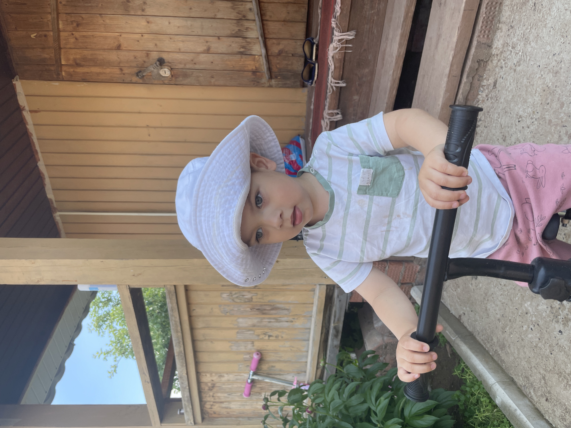 A young child wearing a white sun hat and a striped t-shirt stands in front of a wooden building. The child is holding onto the handlebars of a black scooter. The building has a porch with wooden steps. There is a pink scooter leaning against the fence to the left of the frame, and some green foliage in the foreground. The scene is set outdoors on a bright day. It is the city of Zav'yalovo, Russia.