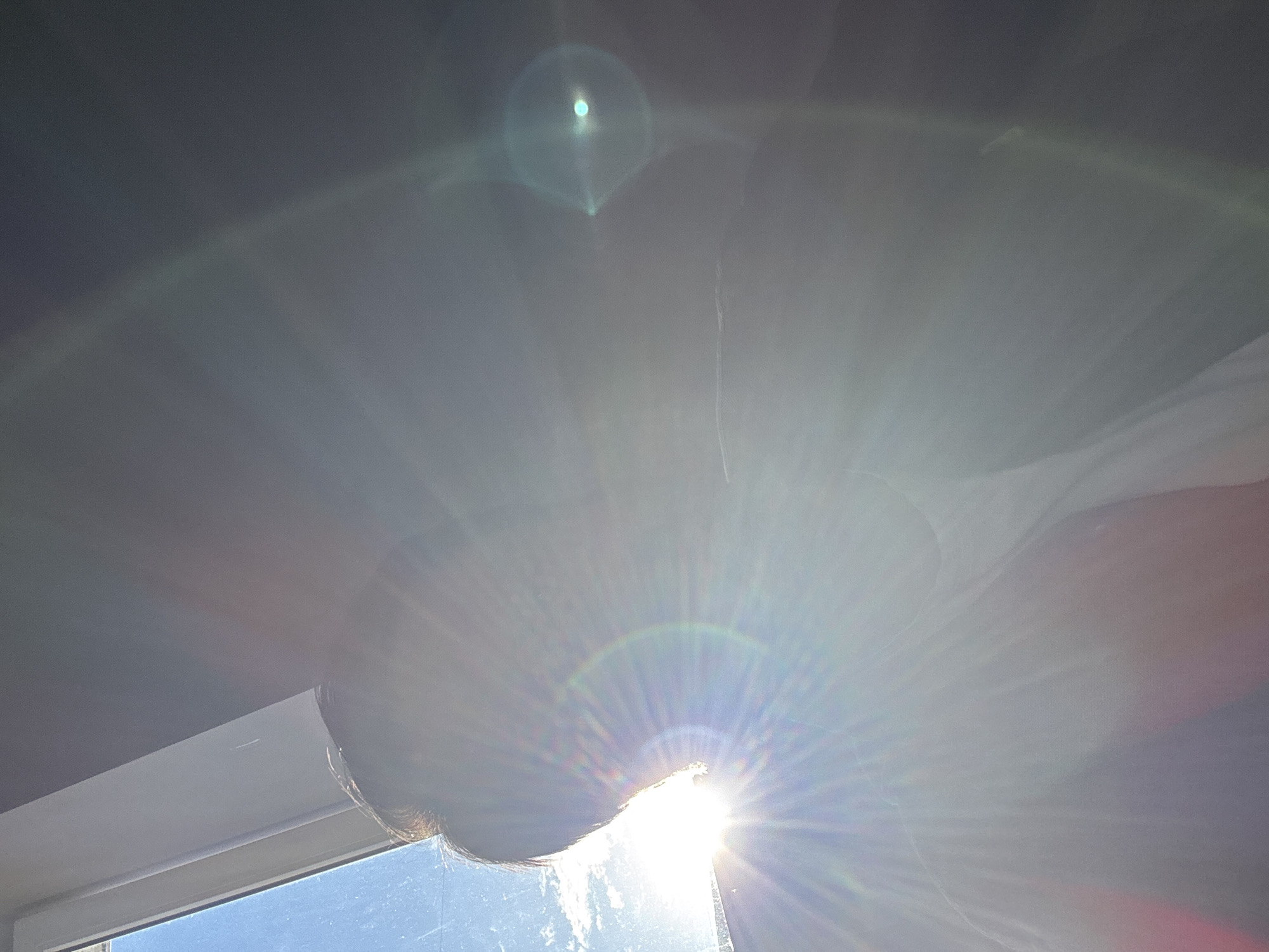 The image shows a view from inside a room, looking out through a window. The sun is shining brightly, creating lens flare and obscuring details of the outside view. The window frame is visible at the bottom left of the image. The upper portion of the image is dominated by a dark area, likely the ceiling of the room. Due to the brightness and glare, it's impossible to identify the specific location in Moscow or any other details of the environment outside the window. No people, subjects, objects or actions are visible in the image.