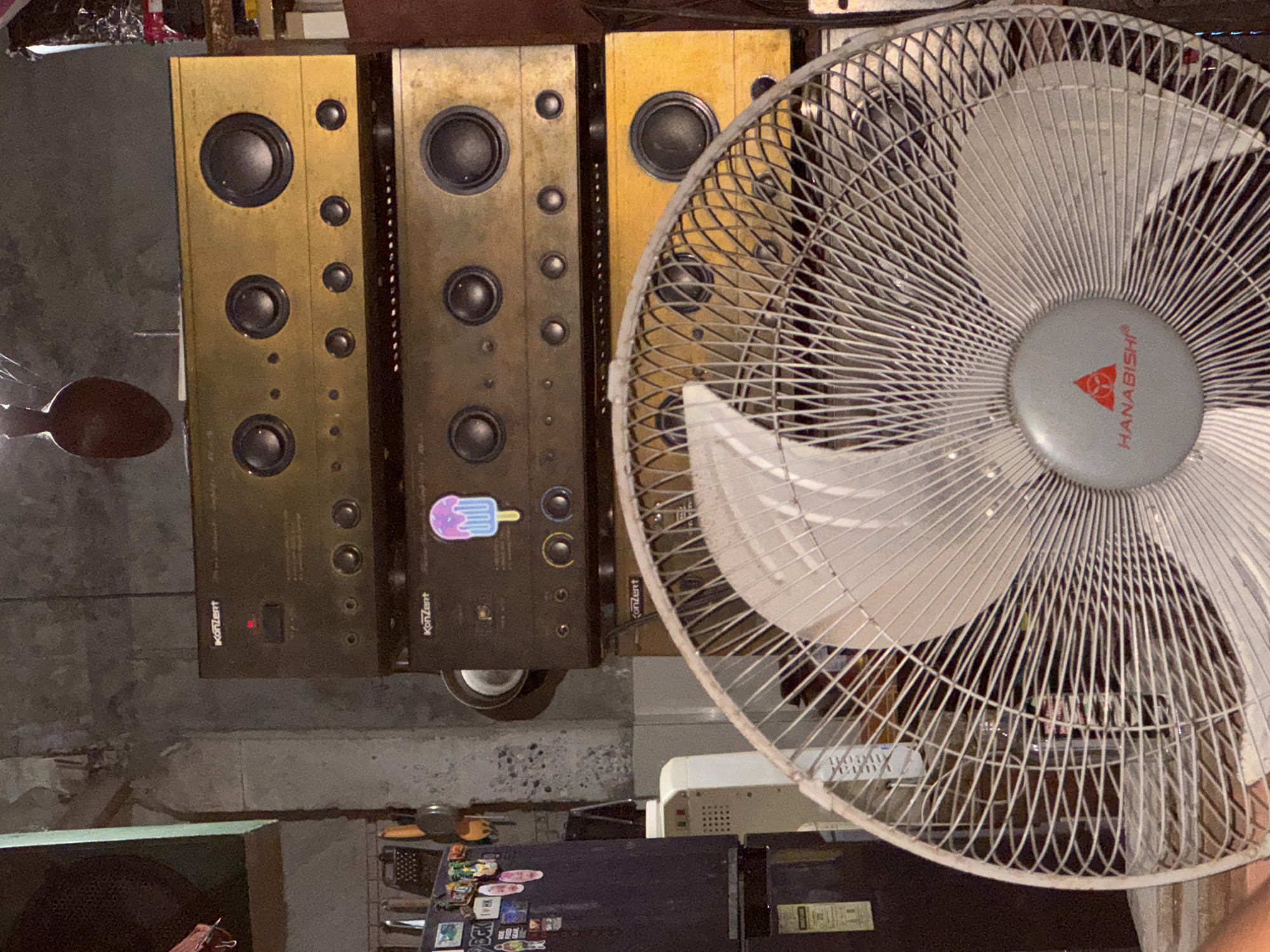 The image shows a stack of three vintage audio amplifiers, likely home stereo receivers, placed on top of each other. The top and bottom amplifiers appear to be gold-colored, while the middle one is darker, possibly black or dark brown. All three amplifiers have prominent circular knobs and speaker output connections on their front panels. The top amplifier has a red indicator light and a label that reads "Konzert." The middle amplifier also has a "Konzert" label and a colorful sticker resembling an ice pop. The amplifiers are stacked in a room with a concrete wall in the background, showing some signs of wear and tear. To the left of the amplifiers, part of a dark-colored appliance, possibly a refrigerator, is visible, adorned with various stickers. To the right and in the foreground, a white oscillating fan with a metal grill and a gray center hub bearing the "HANABISHI" logo is prominently featured. The overall impression is that of a cluttered storage area or a room where audio equipment is kept. The image's context as being located in Davao City, Philippines, suggests that the scene depicts a residential or commercial space within that specific urban environment. There are no visible people or ongoing actions in the frame.