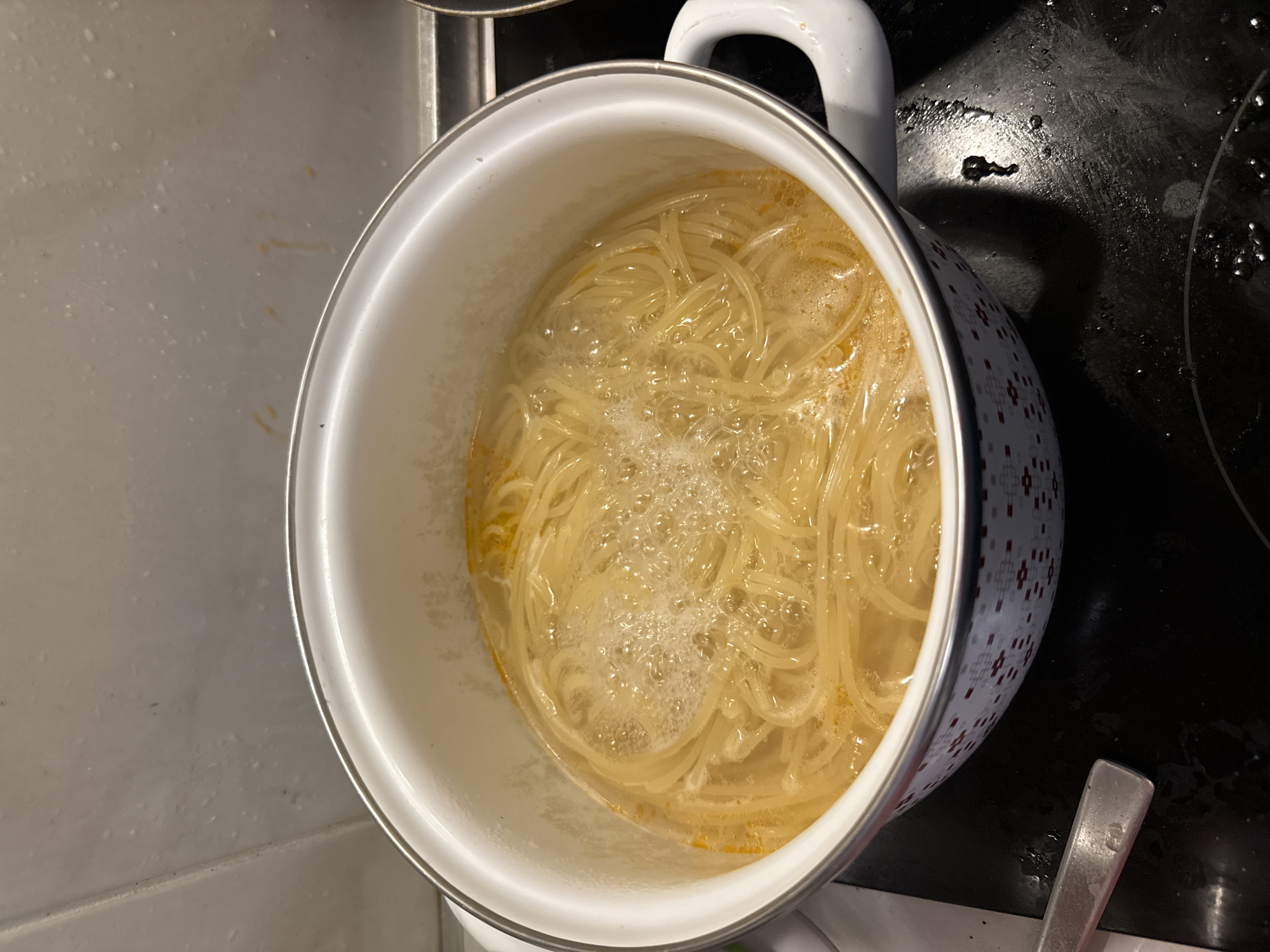 The image shows a close-up view of a pot of spaghetti boiling in water on a stovetop. The spaghetti is golden yellow and appears to be thin, possibly angel hair. The water is bubbling vigorously, indicating it is at a rolling boil. A white pot with a decorative rim featuring a red and grey geometric pattern is filled with the spaghetti and water. The pot has white handles. The stovetop appears to be black and has a few water droplets on its surface. In the background, a white tiled wall with some water stains is visible. The scene is likely taking place in a kitchen, possibly in the city of Boreti, Montenegro, though this cannot be definitively confirmed by the image alone. There are no people visible in the image.
