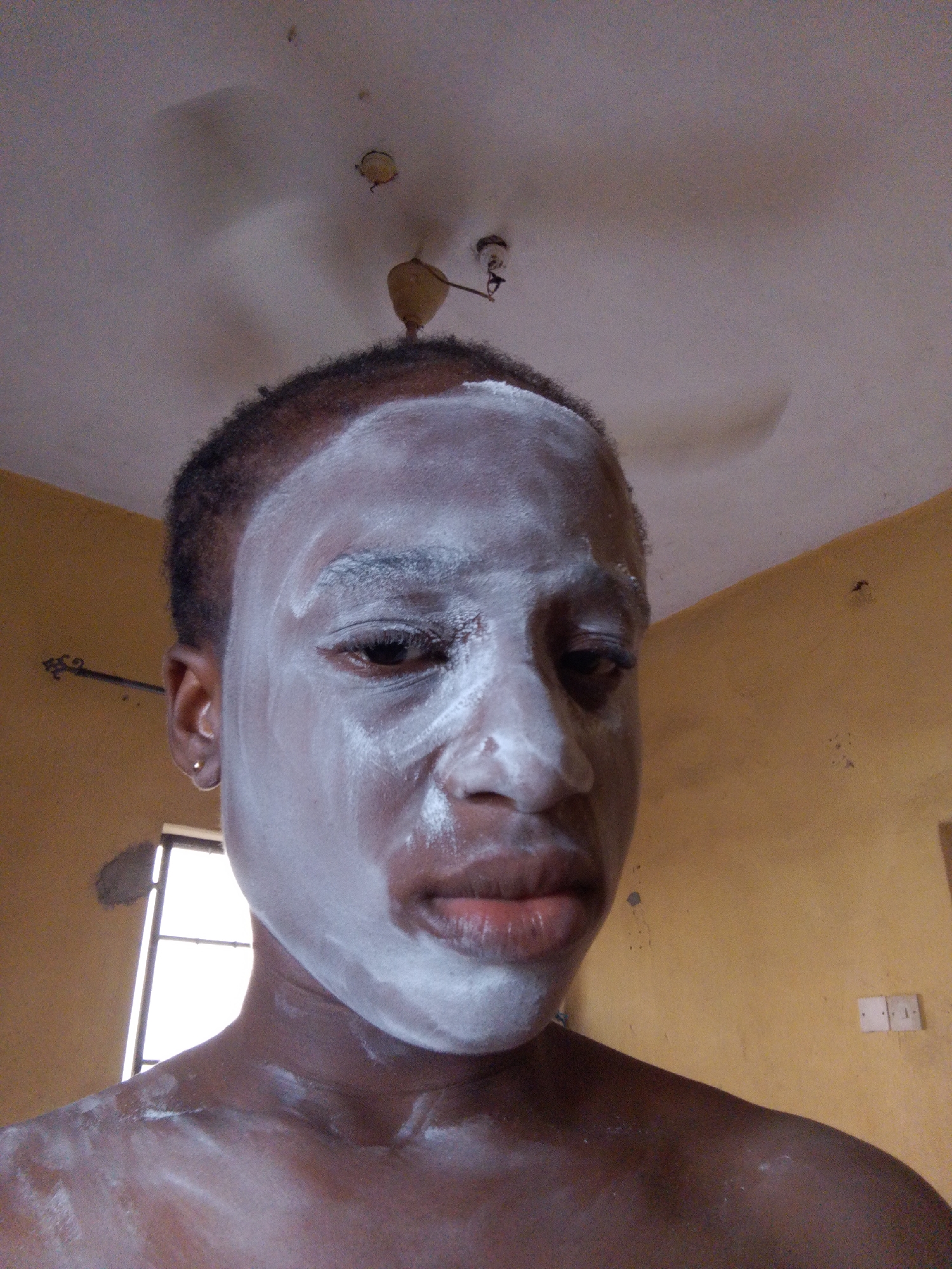 A close-up, upward-angled shot of a person with dark skin wearing a white face mask. The mask covers the forehead, nose, cheeks, and chin. The person's eyes are partially visible beneath the mask, and their lips are slightly parted. A gold stud earring is visible in the person's left ear.
The background consists of a yellow wall with a window in the upper left corner and a white electrical outlet plate on the right. A ceiling fan is visible in the upper center of the image, attached to a white ceiling.
It is not possible to confirm the location as Umuahia, Nigeria without further information.