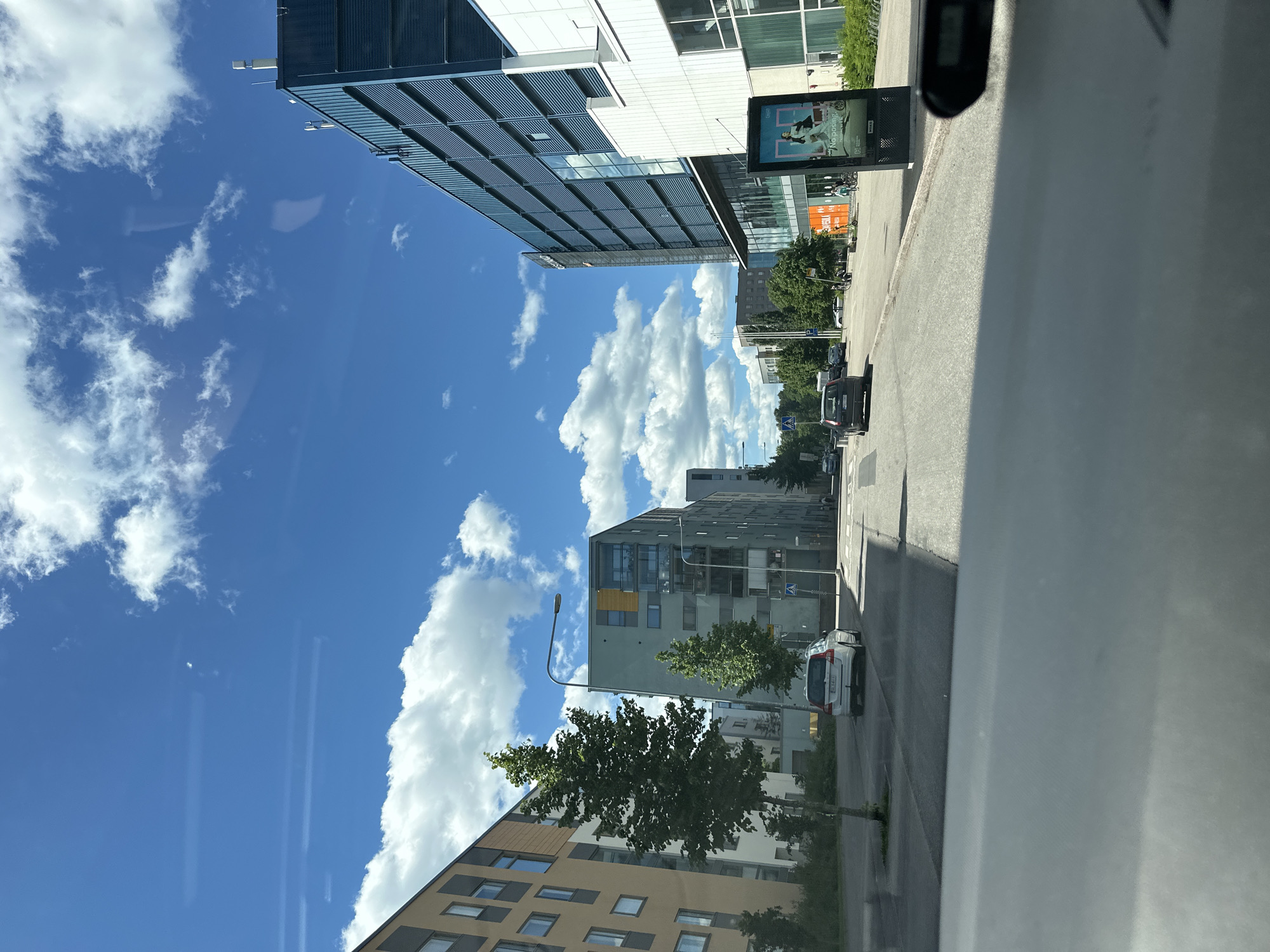The image captures a street scene in a modern urban environment under a clear blue sky with scattered white clouds. On the left side of the street, a contemporary apartment building with a beige facade and dark windows is visible. Adjacent to it, a tall, slim green tree stands prominently. Further down the street on the left, another apartment building with a light green facade and balconies can be seen, with two trees in front of it. A white Volvo station wagon is parked on the side of the road near this building.
On the right side of the street, a large, multi-story building with a dark blue facade and horizontal louvers dominates the view. A white section of the building is visible to its right. In front of this building, a digital advertising billboard displays an image of a person in athletic wear and the text "No posing". Several pedestrians and cyclists are present on the sidewalk in front of this building. Other cars are driving on the street. The foreground of the image shows the interior of a vehicle, with the dashboard and part of the windshield reflected. The scene appears to be a well-maintained, clean urban area.