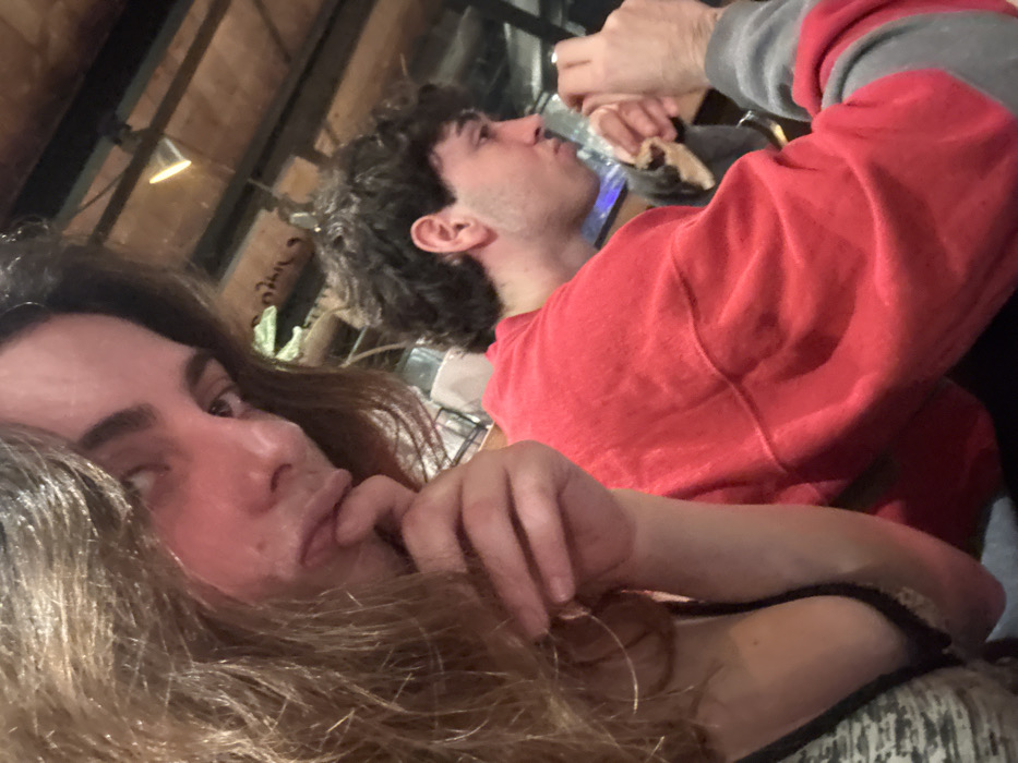 A selfie shows two people. A person with long brown hair gazes at the camera, their fingers near their mouth. In the background, a person with curly dark hair drinks from a clear bottle. They wear a red sweatshirt with gray sleeves. The setting appears to be indoors with wooden beams visible overhead. Other people are partially visible behind the drinking person.