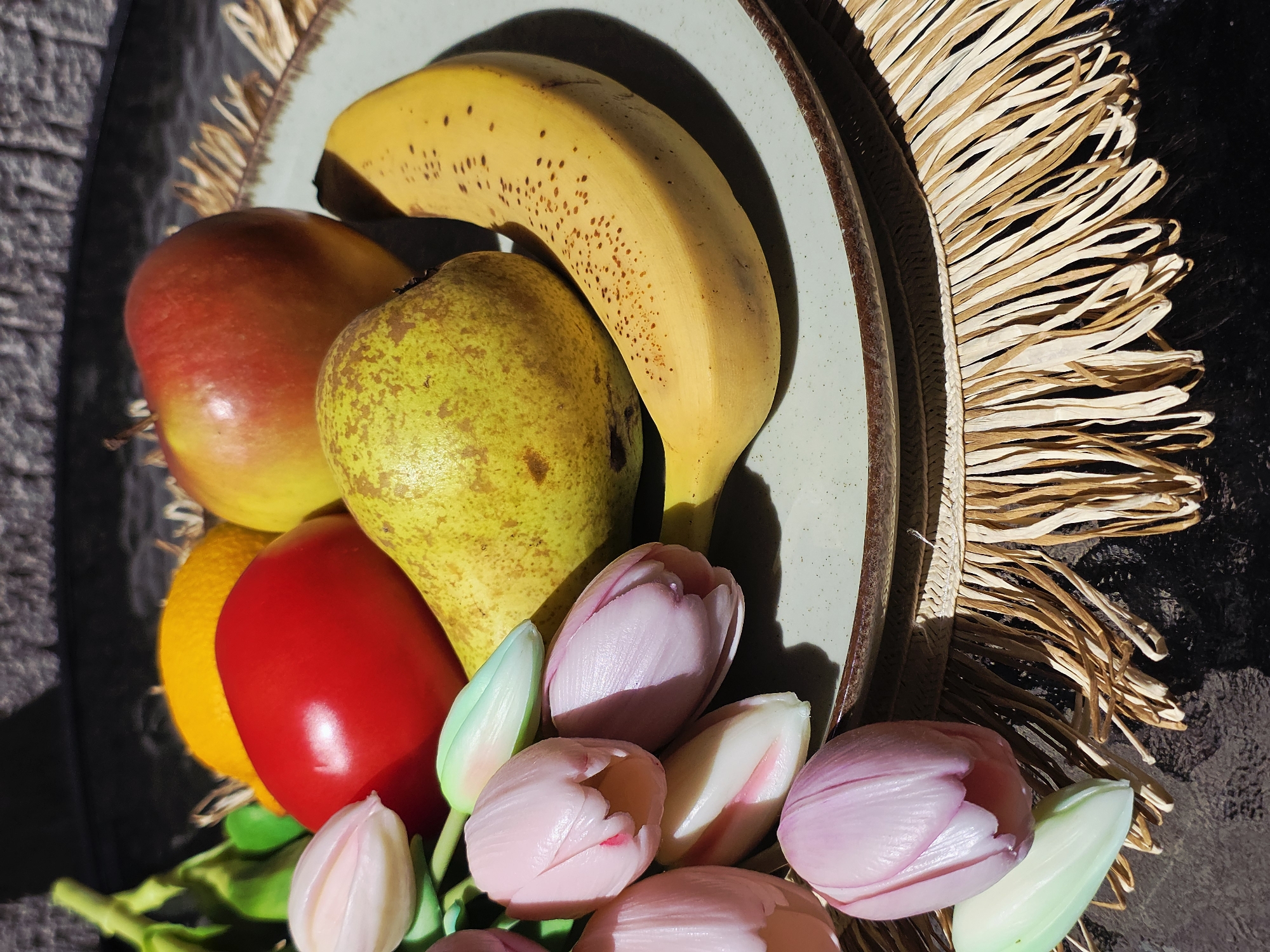 A still life image shows a round plate holding fruit and flowers. The fruit includes a ripe banana with brown spots, a pear, a mango, a red bell pepper, and an orange. The flowers are pink and white tulips. The plate is on a table with a dark surface. A fringed placemat is under part of the plate. The location is unknown.