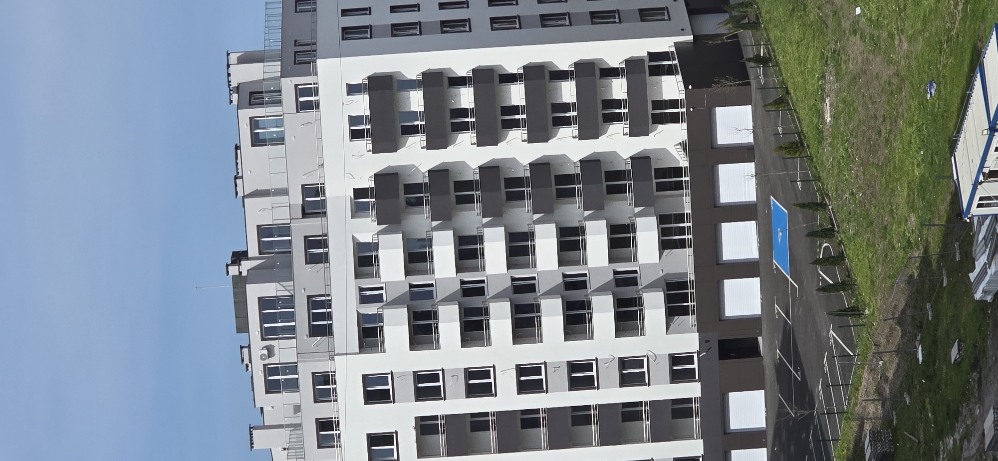 The image captures a modern, multi-story residential building under a clear blue sky, likely in Burgas, Bulgaria. The building features a repetitive architectural style with light grey and white facades, accented by dark grey elements around numerous windows and projecting balconies. Scaffolding is visible on the left side of the building, suggesting it is either newly constructed or undergoing maintenance.
To the right of the building, there is a vibrant green lawn area. Adjacent to the lawn, a dark grey paved surface includes white markings that delineate spaces, possibly parking spots, and a prominent bright blue rectangular section, indicative of a small sports court or play area. A neat row of small, conical evergreen trees borders the paved area, providing a touch of greenery. Further right on the grassy expanse, a small, portable structure with white and blue siding is visible.
The scene is brightly illuminated by direct sunlight, casting distinct shadows from the building's architectural features and the trees, suggesting a clear, sunny day, likely during the morning or early afternoon. There are no people visible in the image, and no specific activities are taking place. No visible text is present within the frame.