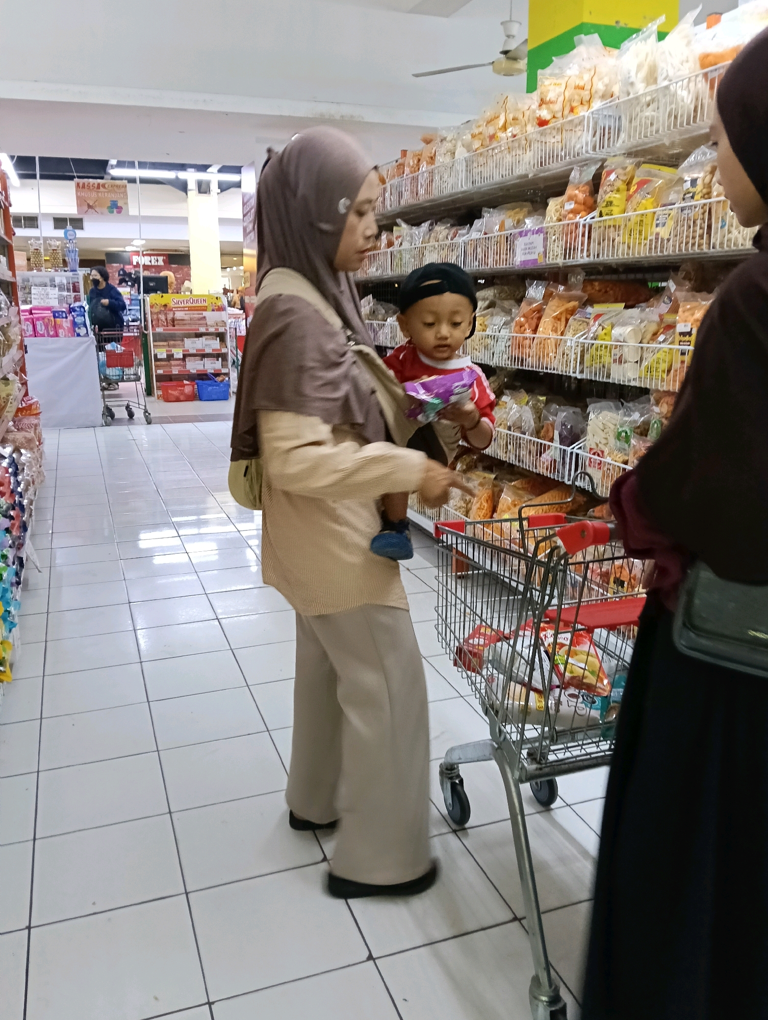 A full-shot, eye-level, outdoor photograph captured during the day shows a woman in a brown hijab and beige clothing standing in a supermarket aisle in Jaten, Indonesia. She is carrying a young child, who is wearing a red shirt, black cap, and blue shoes, in a baby carrier on her chest. The child is holding a small, colorful package in their hands. The woman is pushing a metal shopping cart filled with various food items. In the background, shelves are stocked with packaged snacks. Another person, wearing a dark hijab and black clothing, is partially visible on the right side of the frame. The floor is tiled with white squares and dark grout.