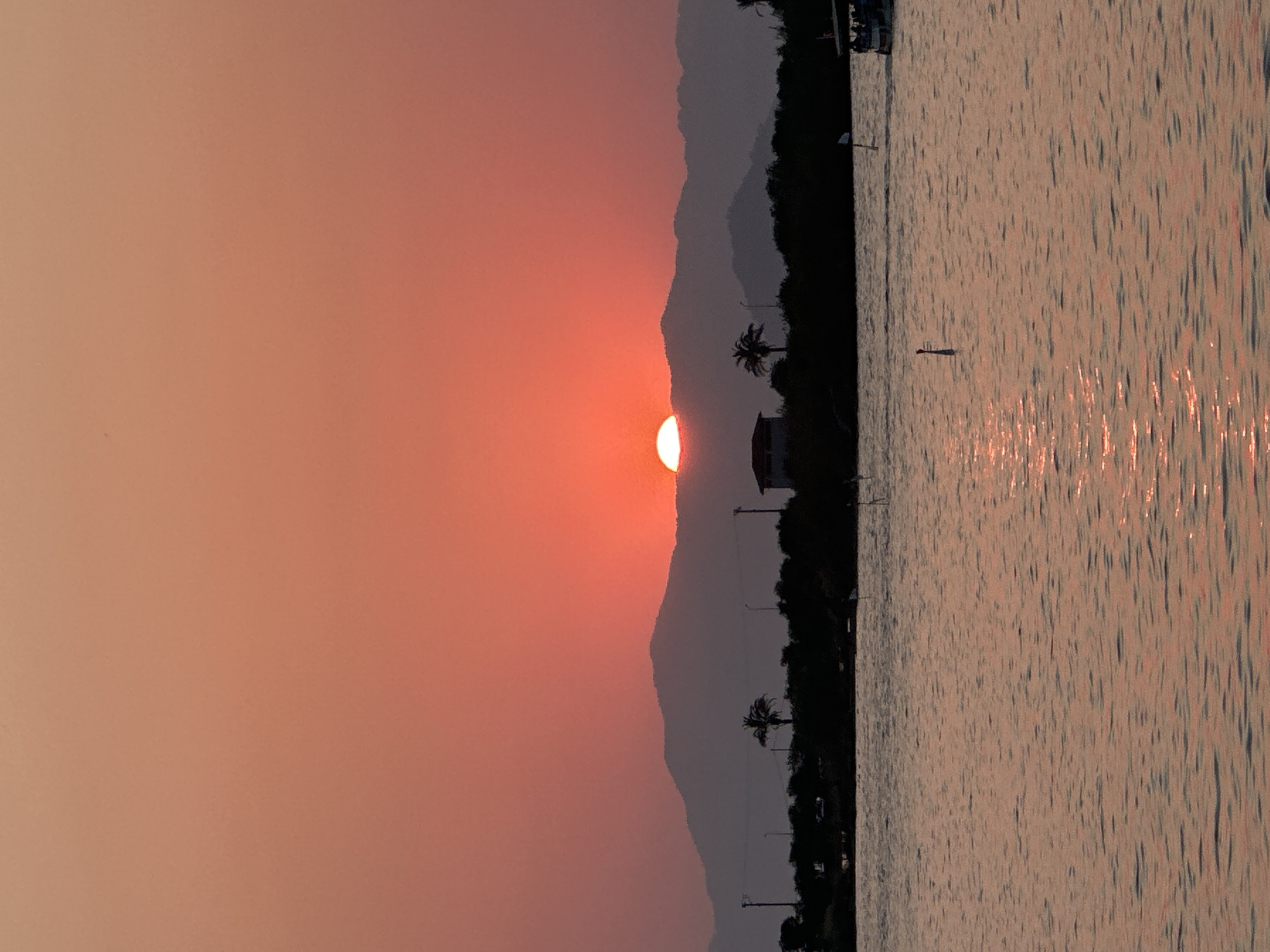 A vertical, full-frame, eye-level shot depicts a tranquil sunset over the water in Fethiye, Turkey. The sun, a vibrant orange disk, is partially obscured by a dark, silhouetted mountain range, casting a warm glow across the sky, which transitions from a soft pink to a deeper orange at the horizon. In the foreground, the calm water reflects the sky's hues and the sunlight, creating shimmering streaks. A dark, tree-lined shoreline runs horizontally across the middle ground, punctuated by the silhouettes of palm trees and a small building. A boat with several passengers is visible on the right side of the frame, and a solitary marker pole rises from the water to the left of the boat. The scene conveys a sense of peace and natural beauty.