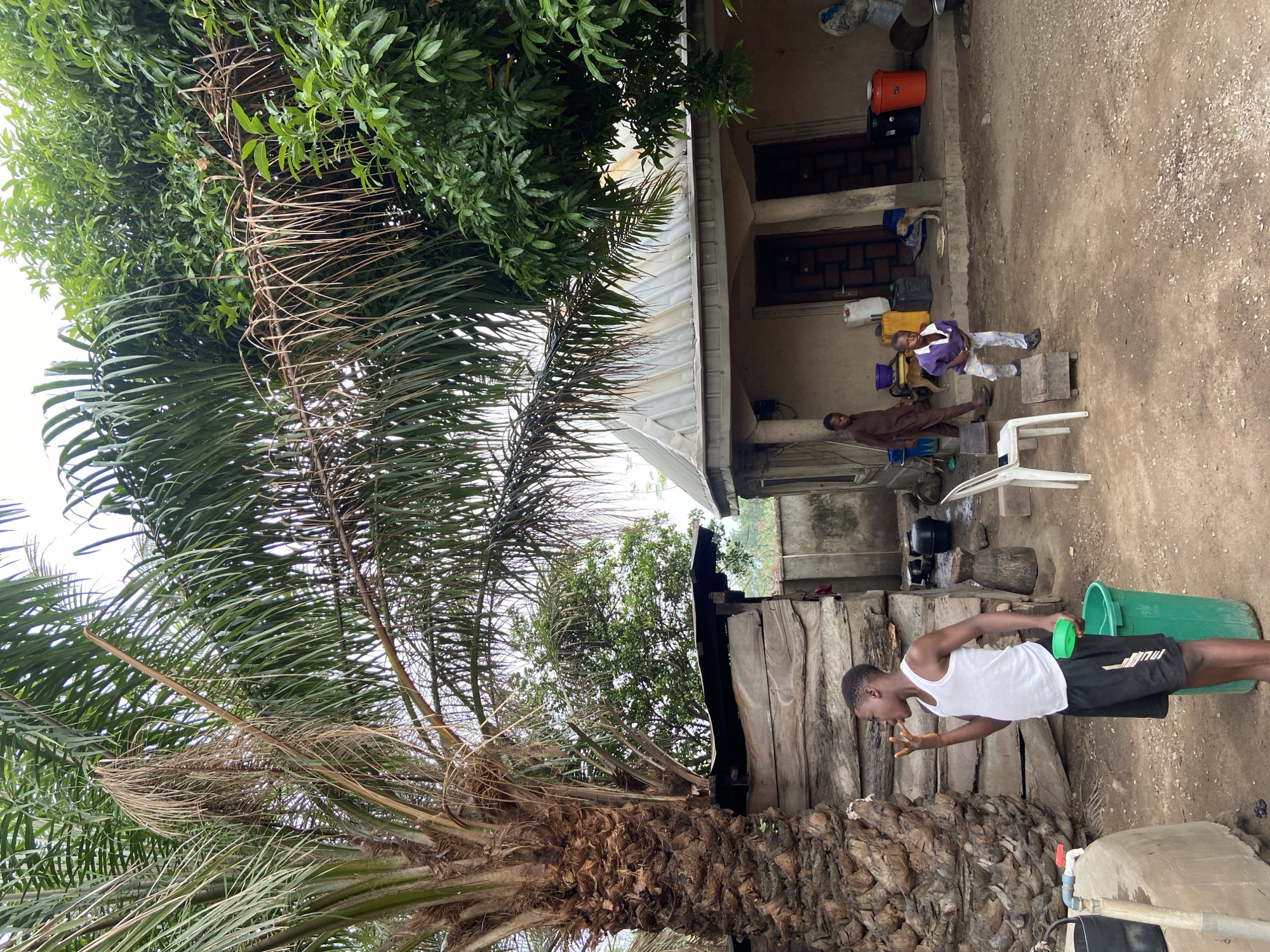 The scene depicts an outdoor area near a building in Mararaba, Nigeria. A young man in a white tank top and black shorts is drinking water from his hands while holding a green bucket. Nearby, a building with a metal roof has two people sitting under its covered porch: one in brown attire and another in purple, both sitting on chairs and benches. A white plastic chair sits near them. Various objects, including water containers and cooking pots, are scattered around the porch. Vegetation, including a palm tree and other leafy trees, surrounds the area. The ground appears to be dirt and gravel.