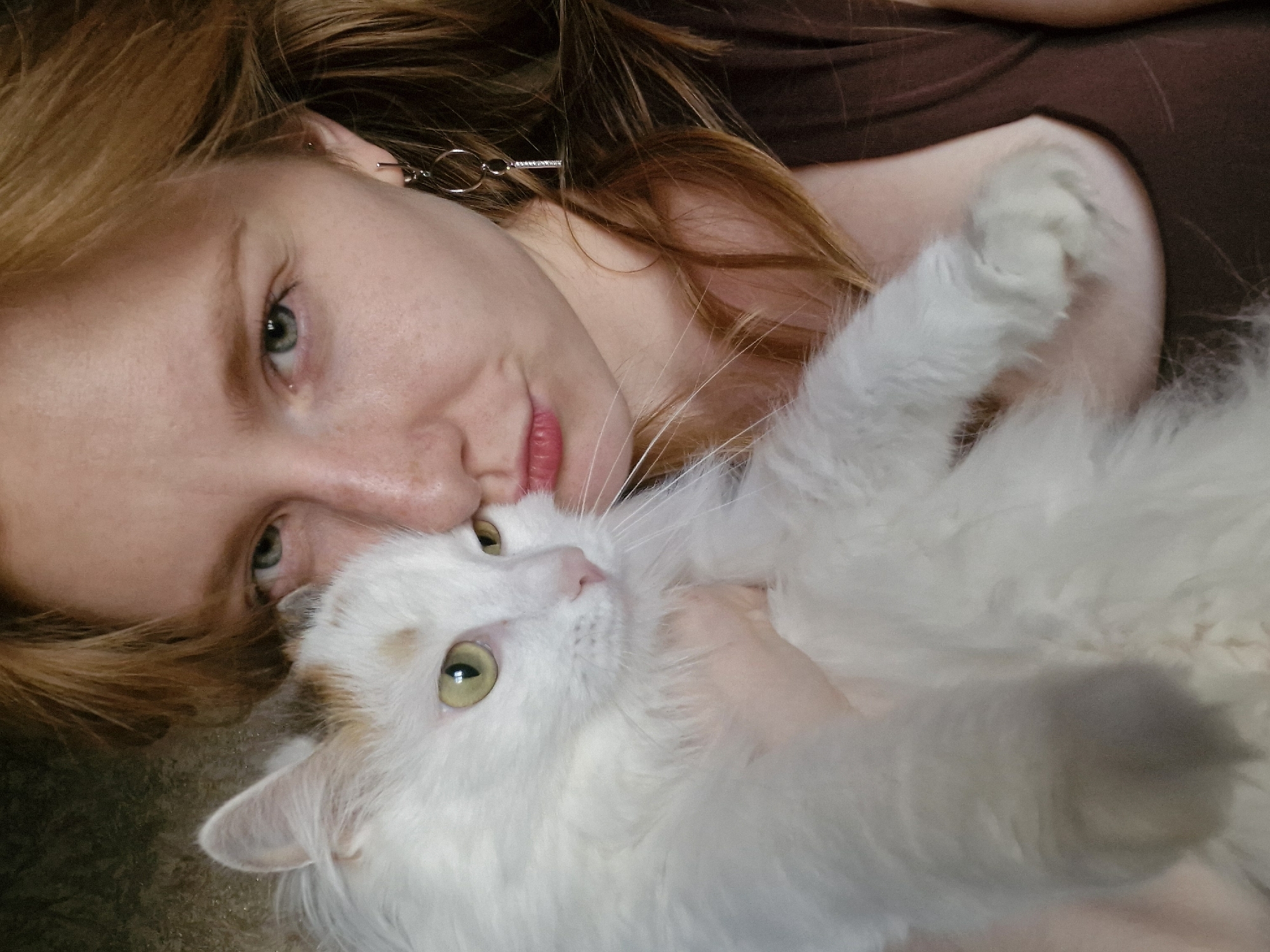 A fair-skinned person with light red hair is lying down, cradling a white cat with light orange markings. The person's face is close to the cat, their noses nearly touching. The cat's eyes are open and looking off to the side. The person is wearing a dark brown top and earrings. The location is not discernible from this image.