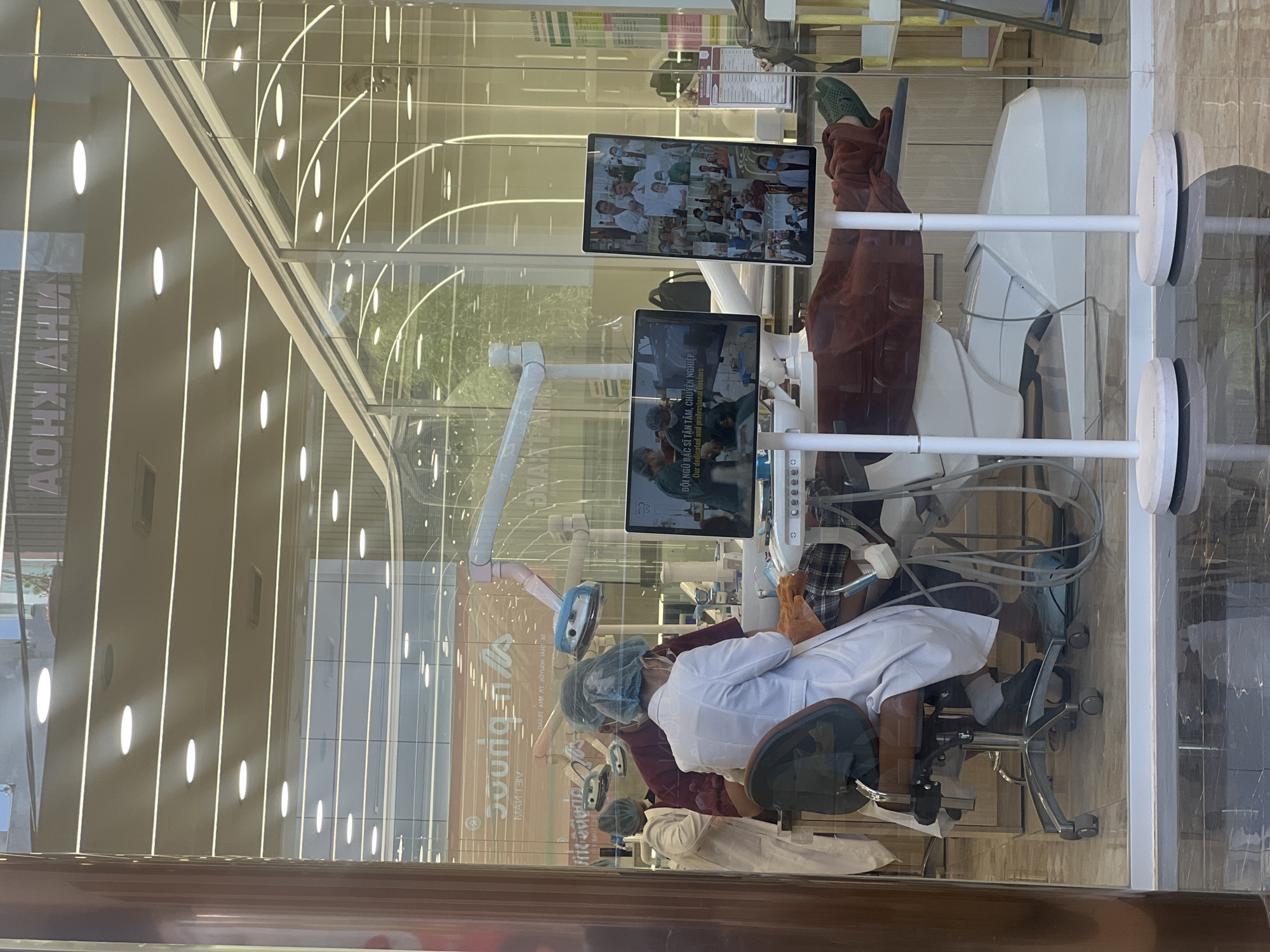 The image shows a dental clinic viewed through a glass window, likely in Nha Trang, Vietnam, given the Vietnamese text on the signage. Inside the clinic, medical professionals wearing surgical caps and white coats attend to a patient in a dental chair. Modern dental equipment is visible, including a monitor displaying medical information. A second monitor shows a collage of smiling medical staff. The interior design of the clinic appears modern, with sleek lighting. The reflections on the glass window suggest the clinic is located in an urban setting.
