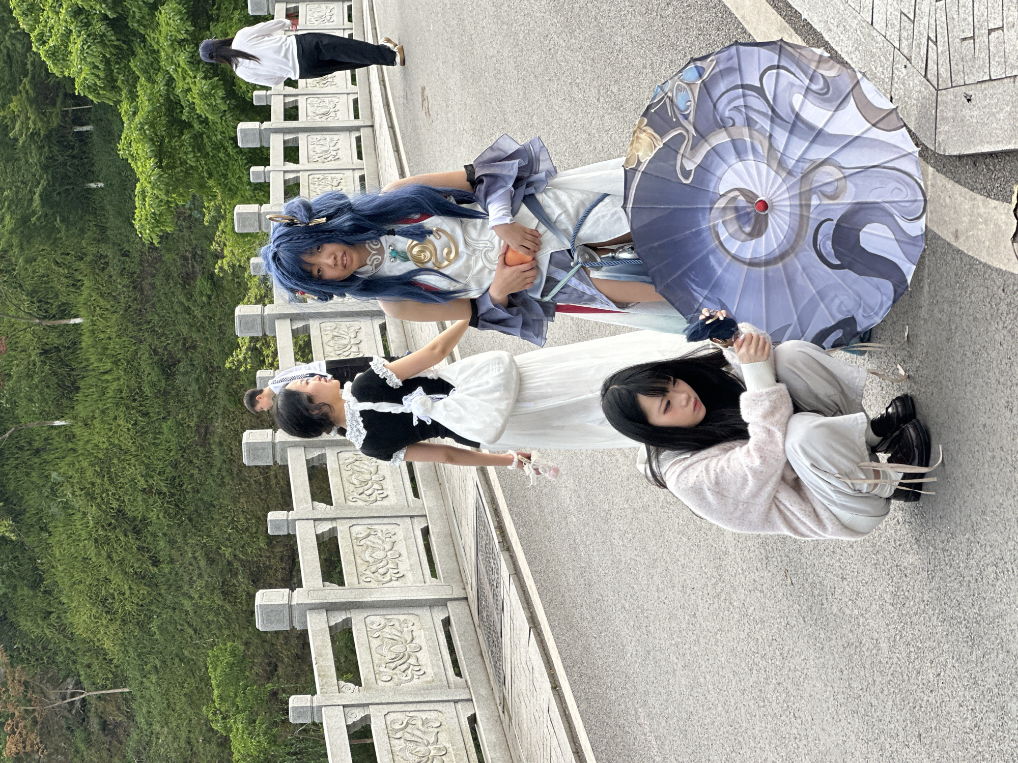 The image shows a group of people posing on a paved area next to a stone balustrade and a green area. One person is dressed in cosplay, with blue hair, a white and blue outfit, and holding an orange. Another person is in a black and white dress. A third person is sitting on the ground, wearing a pink sweater and light pants. An umbrella with a swirling design is near the person sitting. In the background, another person is walking along the balustrade. The location is in the city of Liangjiang, China.