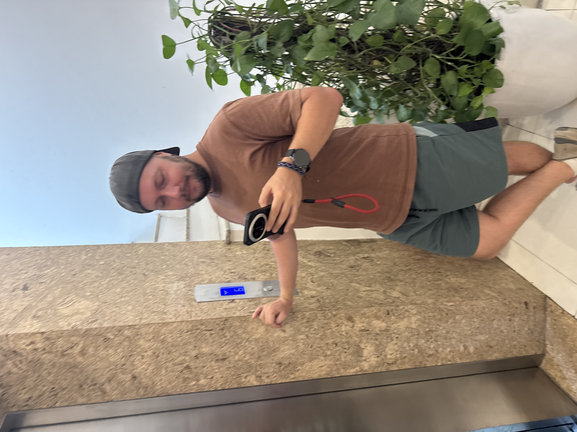 A young man wearing a black baseball cap, a brown t-shirt, and grey shorts is standing by an elevator. He is holding a smartphone and appears to be looking at it. To his right is a large potted plant with green leaves. The elevator panel shows the number "6" lit up in blue. The background consists of a light blue wall and white stairs. The overall scene suggests an indoor setting, possibly a hotel or residential building.