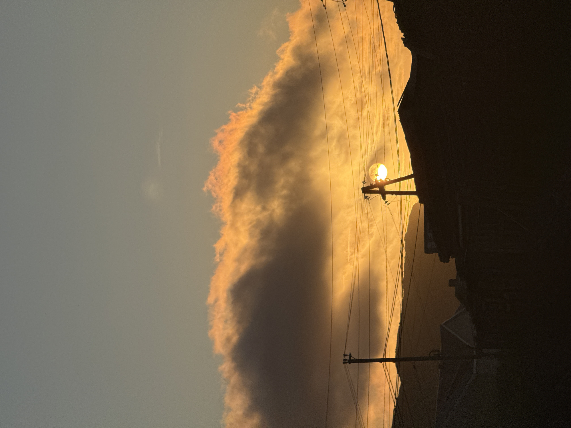 An outdoor, vertical, medium shot captures a sunset in Teriberka, Russia. The sun, a bright orb, is partially obscured by the silhouette of a utility pole and power lines that crisscross the frame. The sky transitions from a pale blue at the top to warm oranges and yellows near the horizon, illuminated by the setting sun. A large cloud formation, tinged with fiery orange along its edges, dominates the upper portion of the sky. Below, the dark silhouettes of houses and buildings create a stark contrast against the vibrant sky, with the mountainous landscape visible in the distant background.